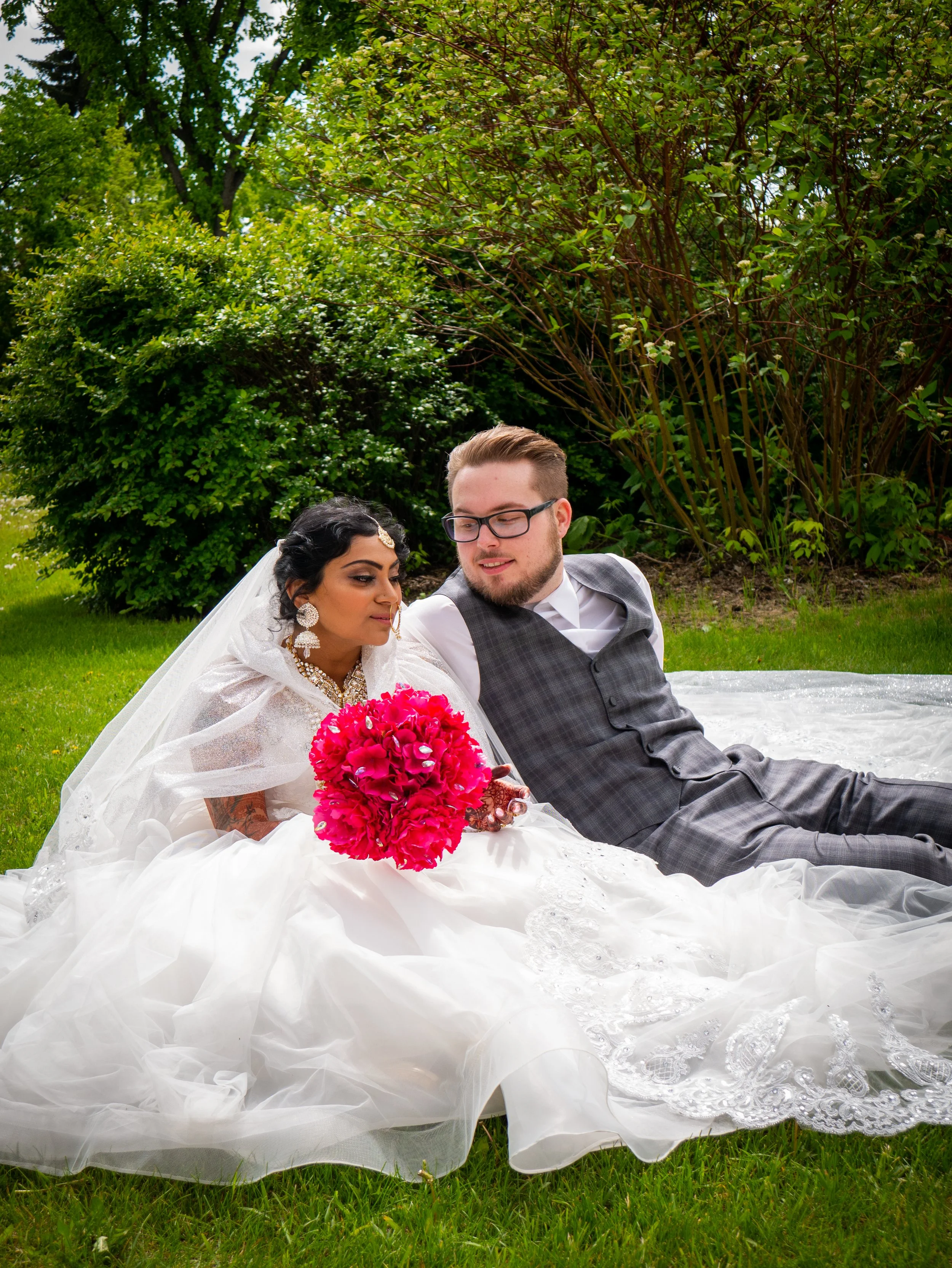 A bride and groom sitting on the grass during a wedding photo shoot outdoors, with lush green trees in the background. The bride wears a white wedding gown with lace and holds a bright pink bouquet, while the groom sports glasses and a gray vest over