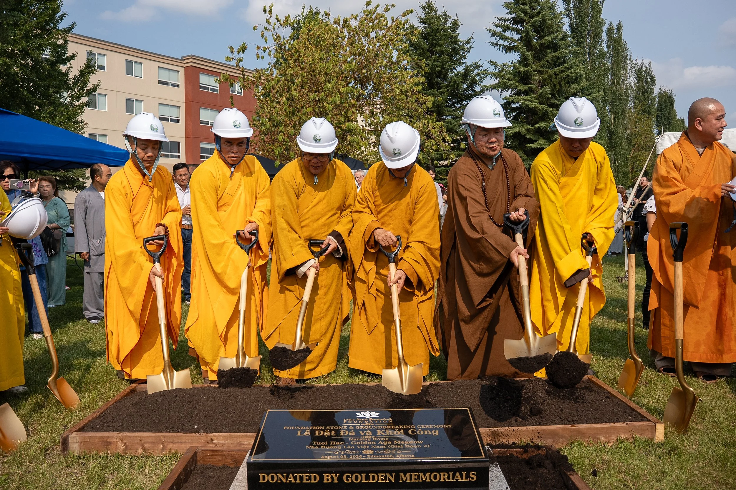 Group of monks and community members participating in a groundbreaking ceremony, wearing yellow and brown robes and white helmets, with shovels in hand, standing behind a wooden-bordered plot of freshly turned soil with a plaque that reads 'Donated b