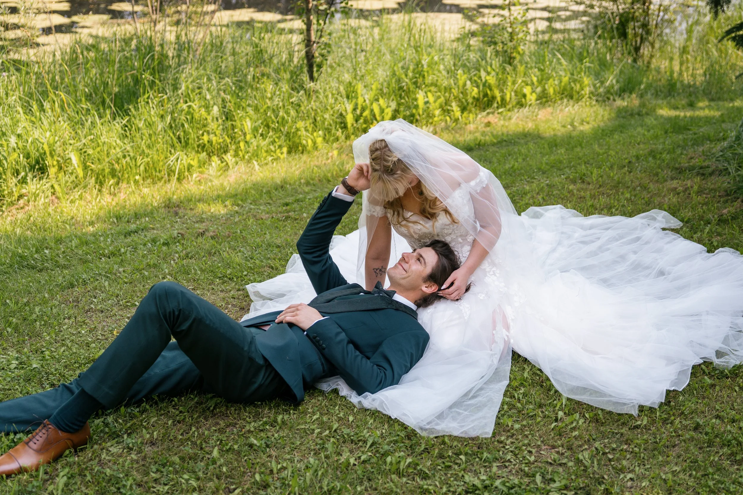A bride and groom lying on the grass in a park. The bride is wearing a wedding dress and veil, leaning over the groom who is dressed in a suit. They are smiling and looking at each other amidst greenery and trees.
