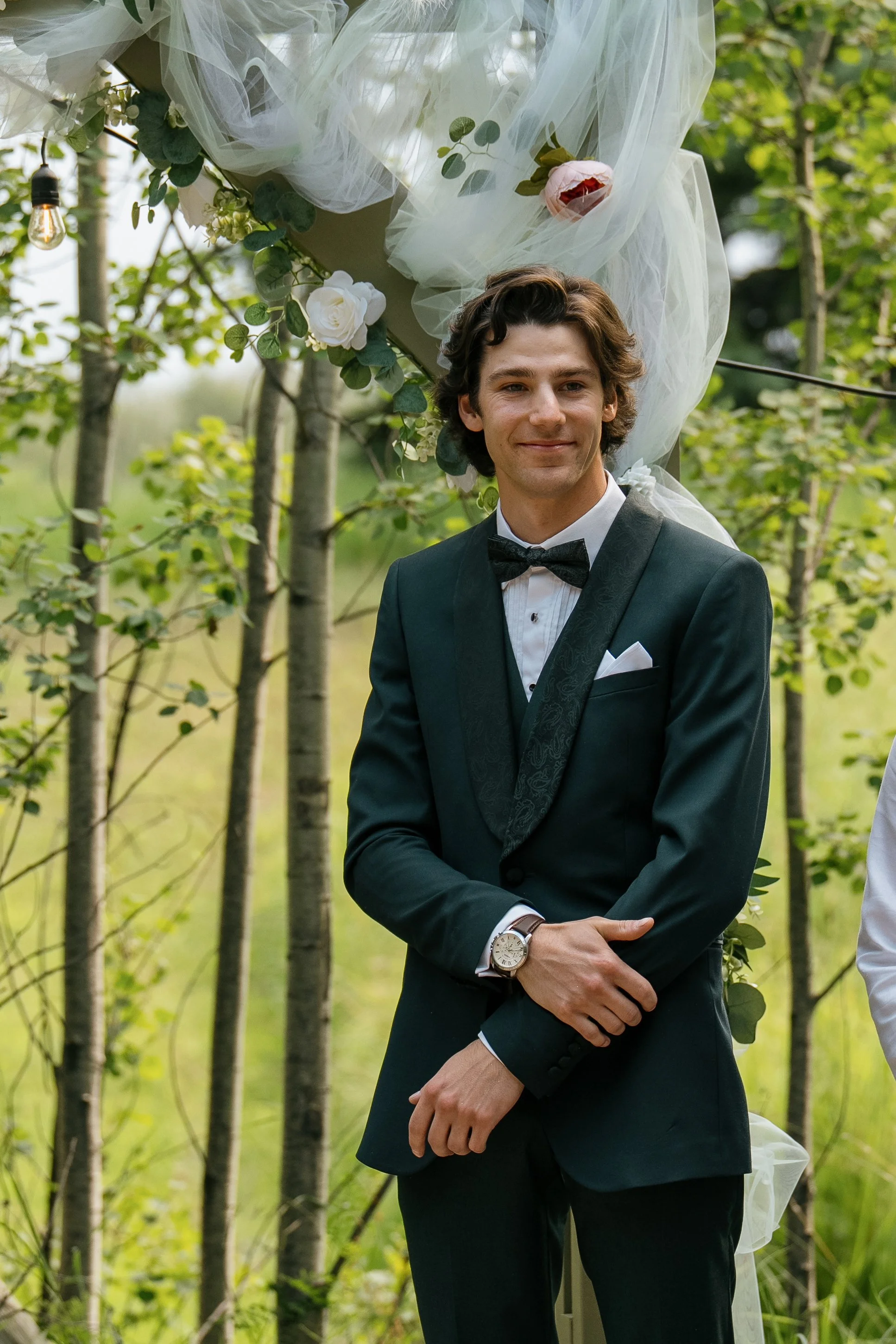 A man in a tuxedo with a bow tie, standing outdoors in front of a decorated arch with flowers and tulle. The setting appears to be a wedding or celebration.