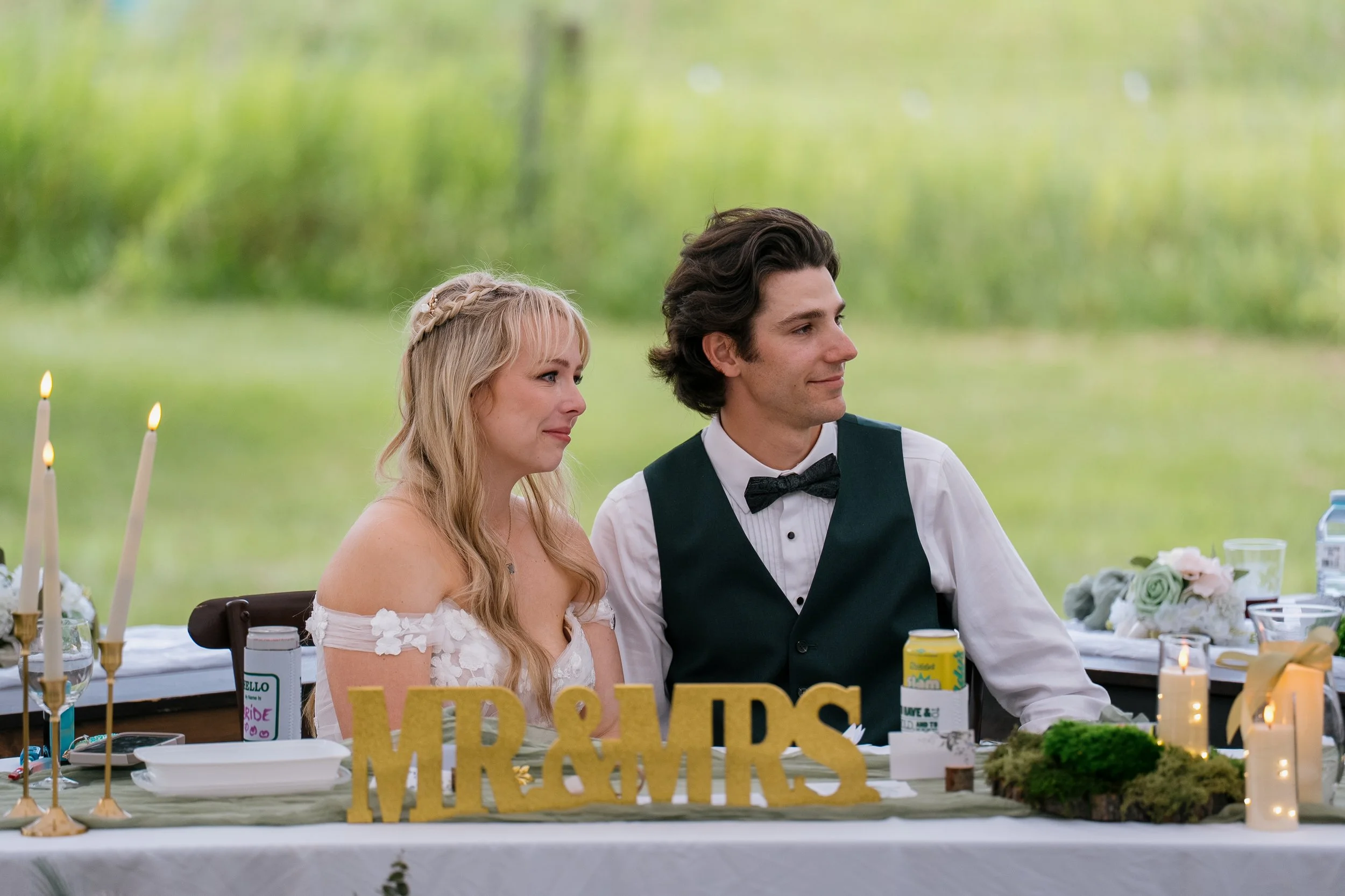 A bride and groom sitting at a wedding reception table outdoors with greenery in the background. The bride has long blonde hair and is wearing a white dress with floral details. The groom has dark wavy hair and is wearing a black vest and bow tie. Th