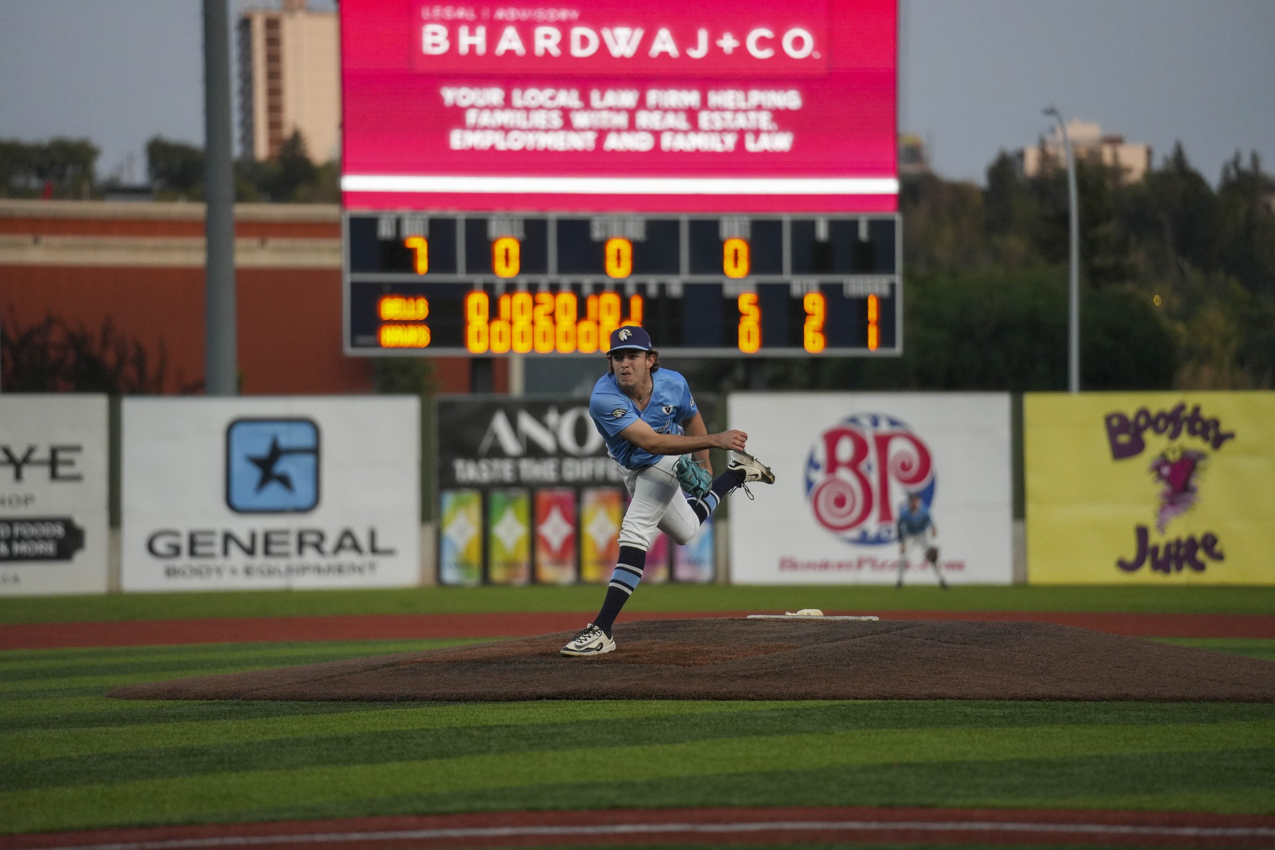 A young baseball player in a blue uniform and cap prepares to pitch on the field, with a digital scoreboard and advertisements in the background.