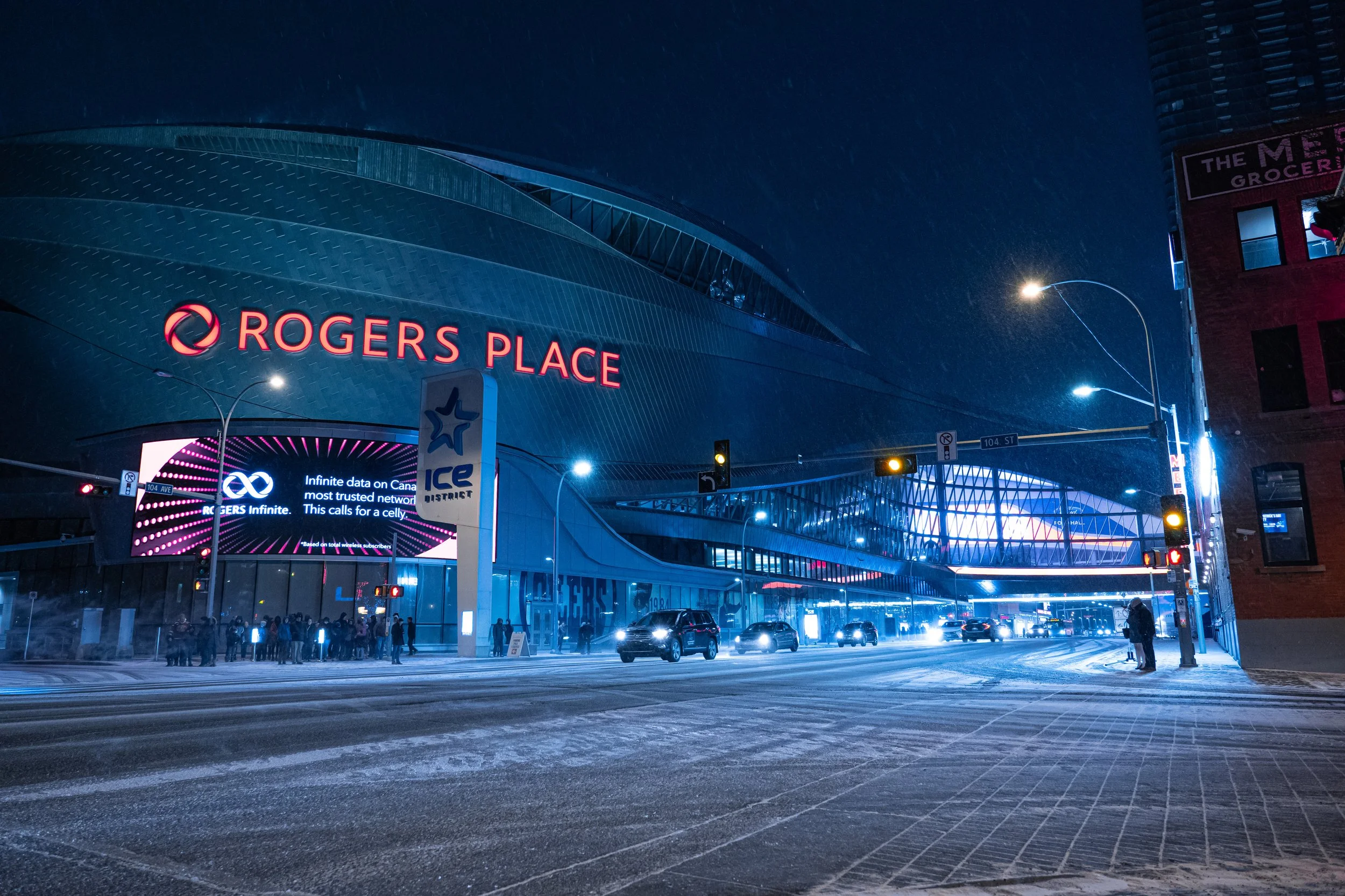 Nighttime view of Rogers Place, an arena with a sleek, modern design, illuminated signs, and a crowd of people outside. Streetlights and traffic signals are visible.