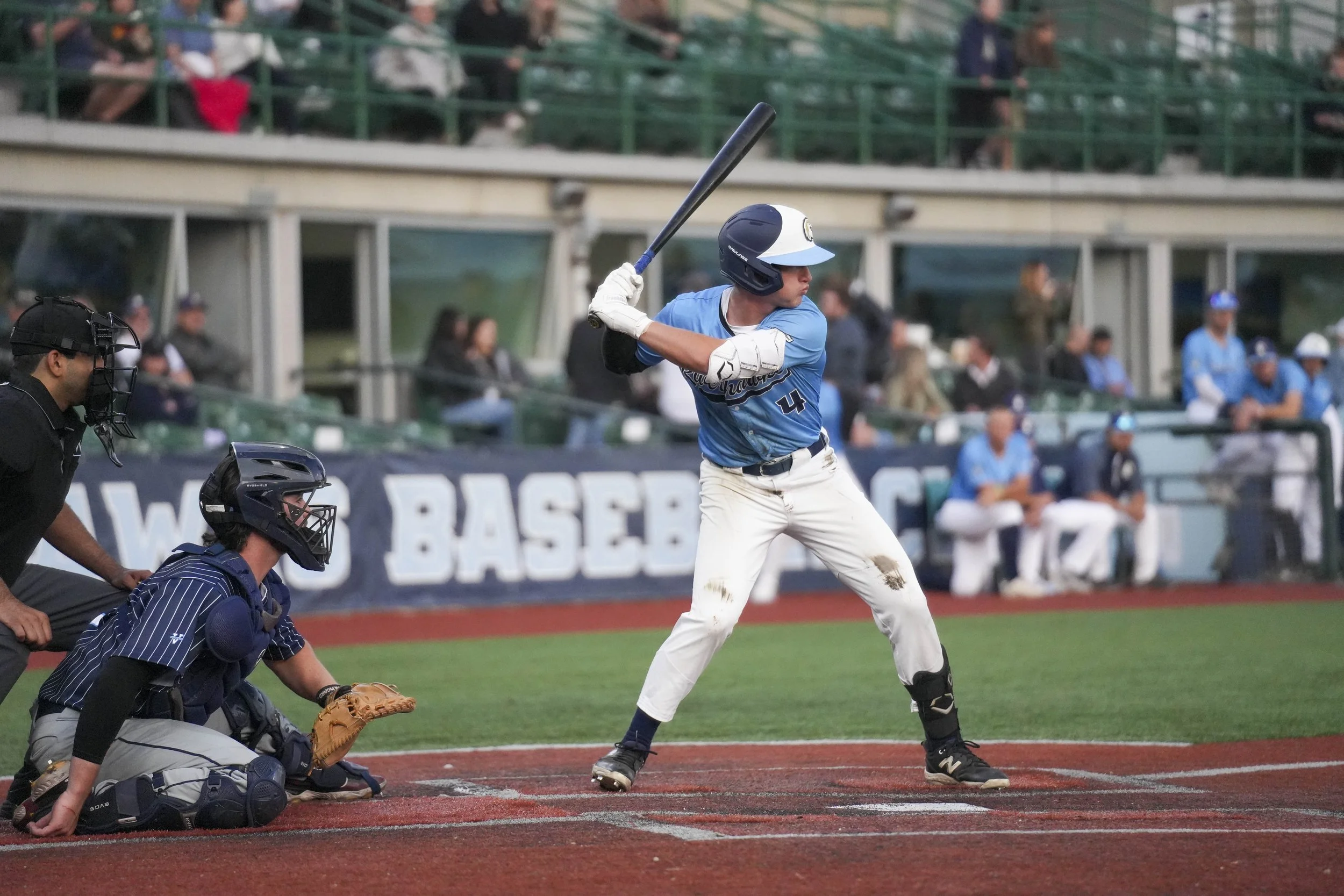 A baseball player in a blue uniform and helmet is swinging a bat at a pitch during a game. An umpire and a catcher are behind him, and spectators are seated in the background.