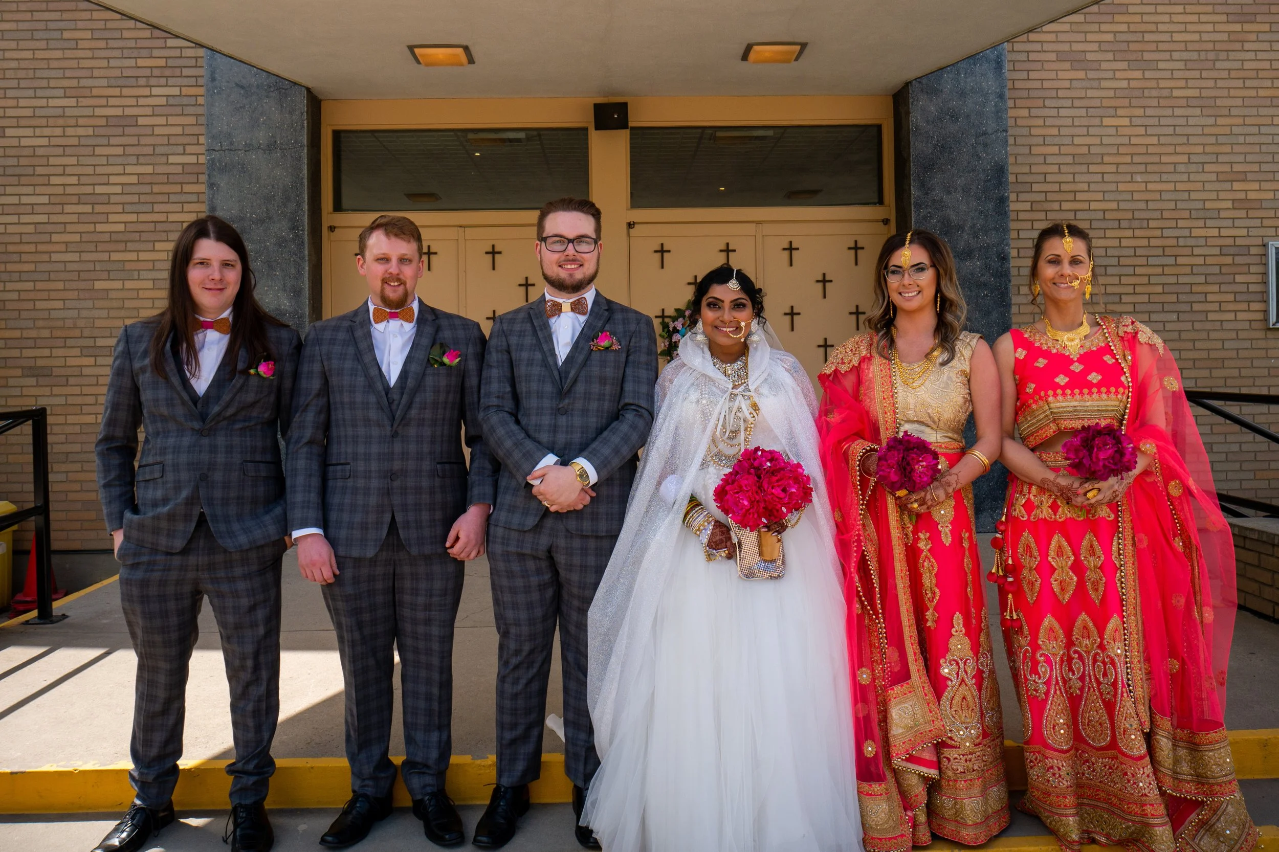 A group of six people dressed in wedding and traditional Indian attire standing outside a church with crosses on the doors, smiling for the camera.