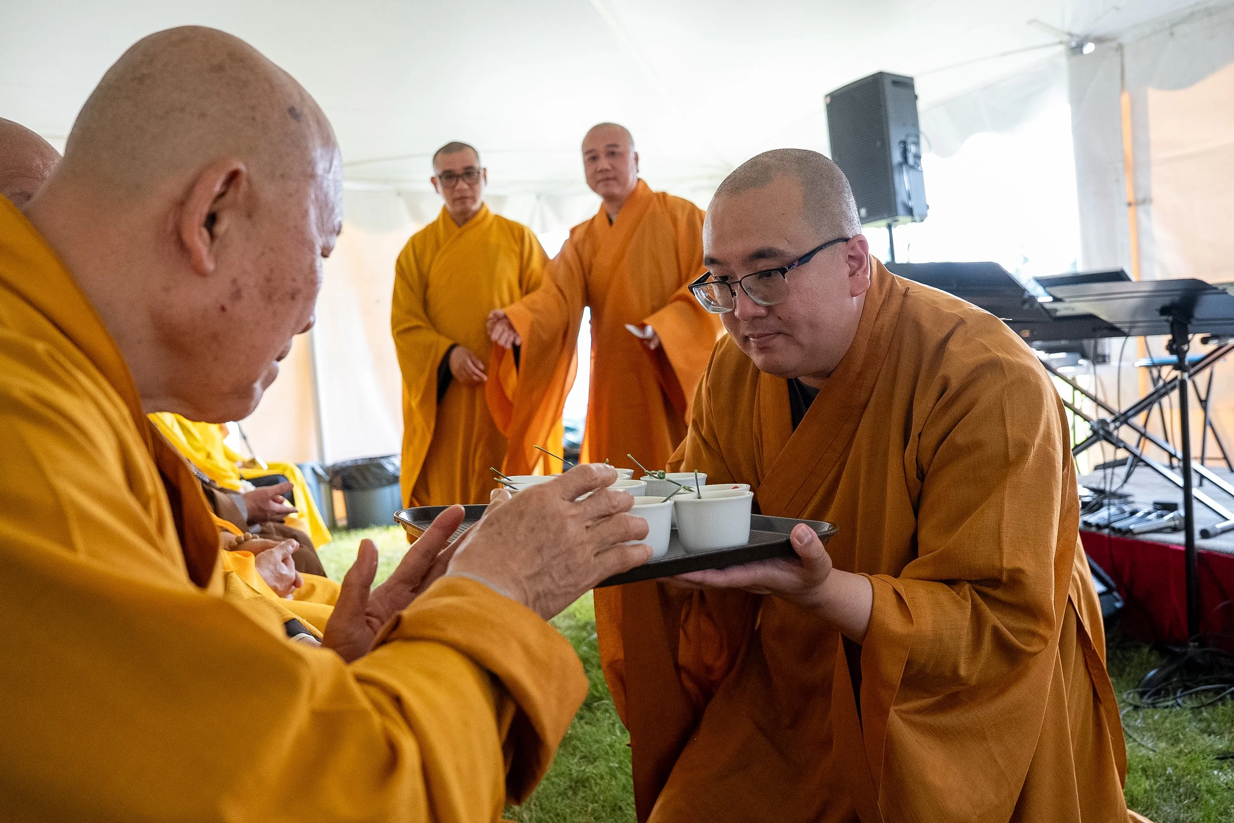 Buddhist monks dressed in orange robes participating in a tea ceremony under a tent, with musical equipment in the background.