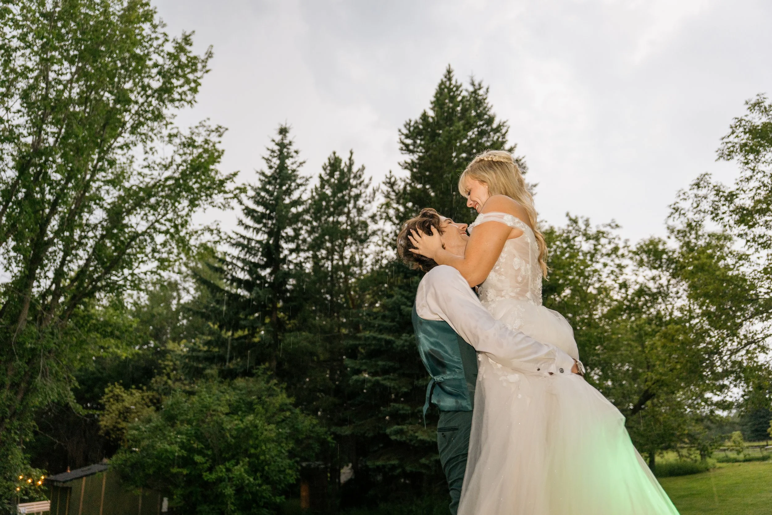 A happy couple dressed in wedding attire, with the groom lifting the bride in his arms outdoors among trees, smiling and sharing an intimate moment.