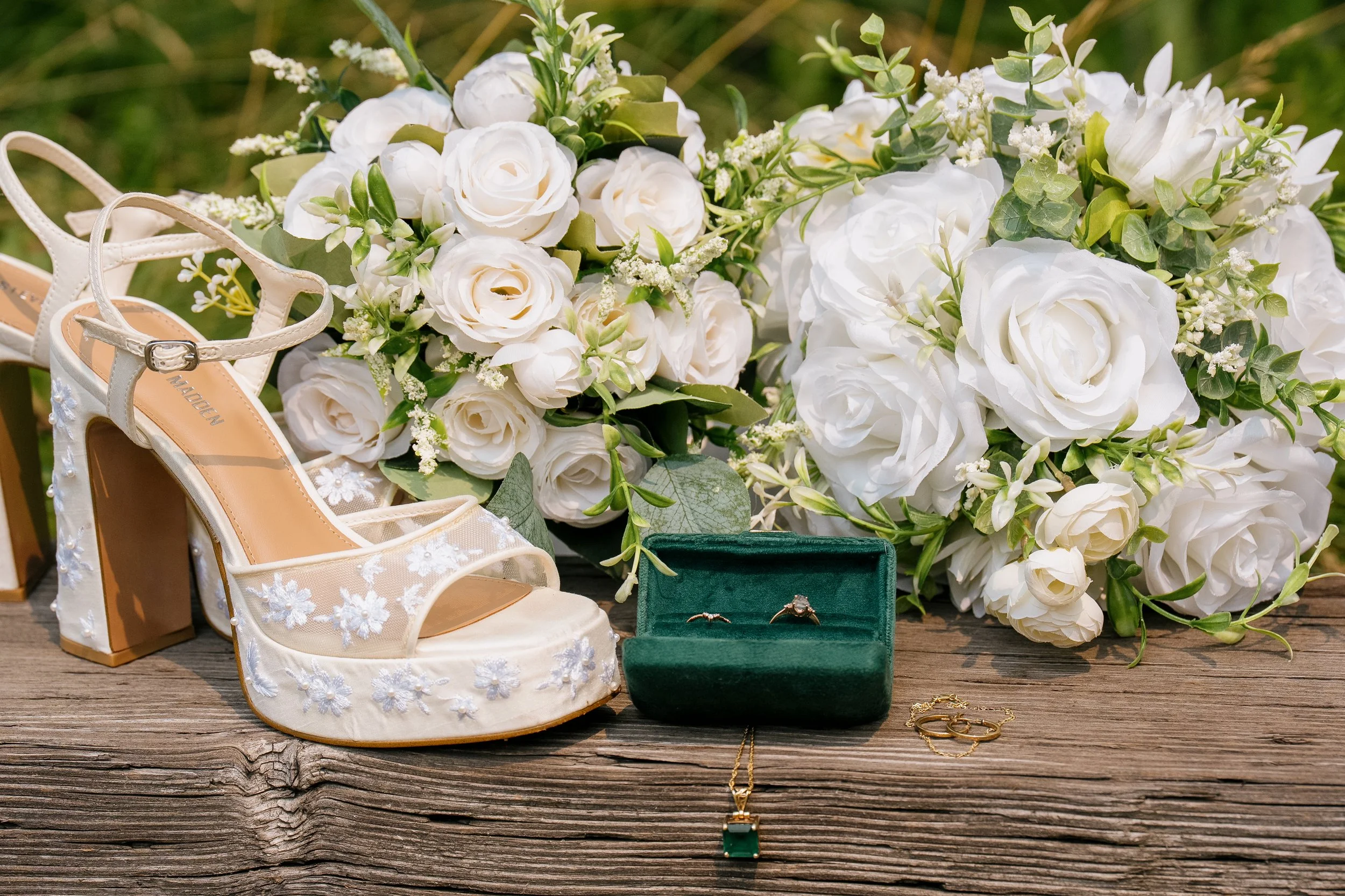 A collection of wedding accessories on a wooden surface, including white bridal shoes with floral embroidery, a bouquet of white flowers, a green jewelry box with rings, and gold jewelry, with a green gift box and a boutonniere in the background.