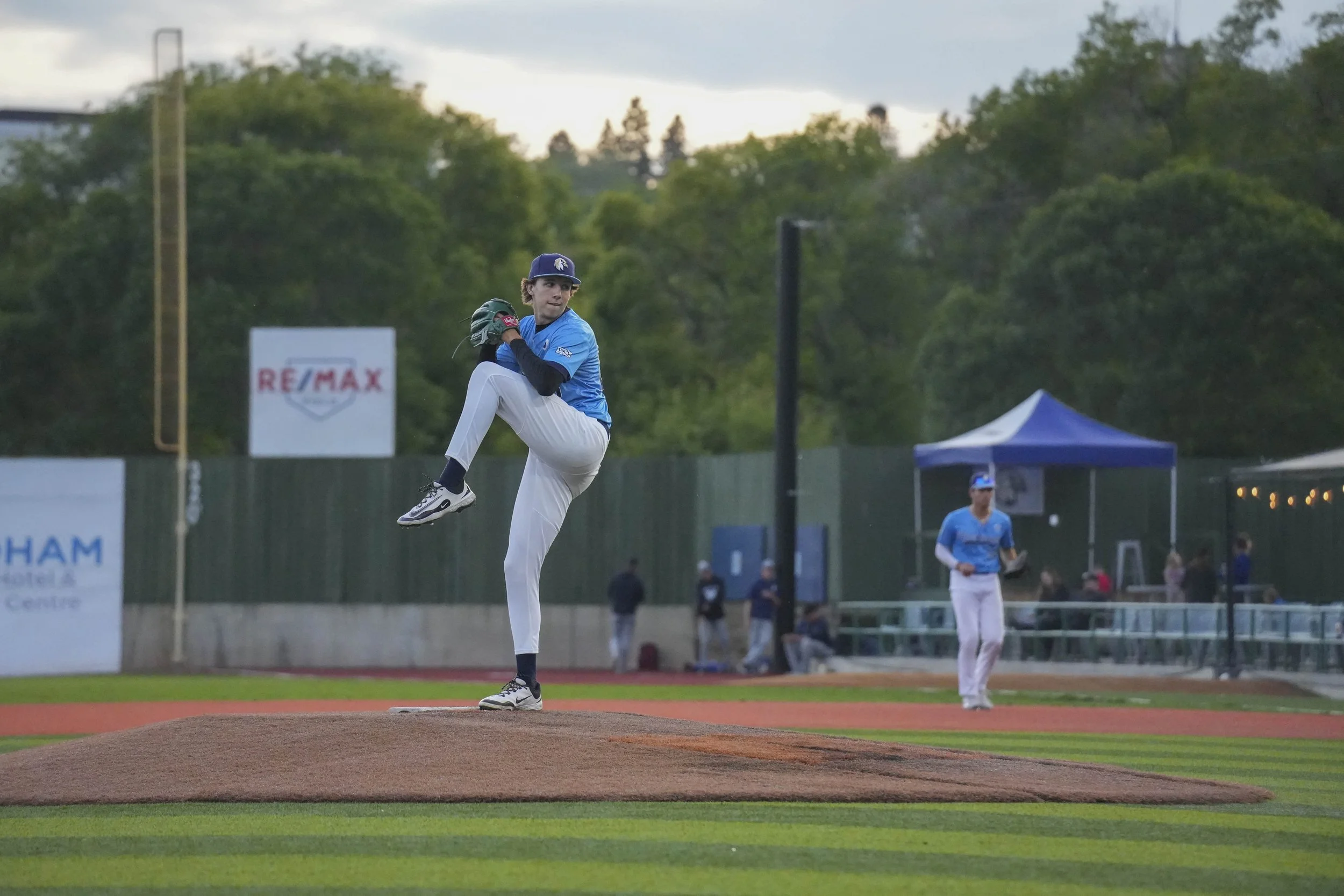 A young baseball pitcher in a blue jersey and white pants is pitching on a baseball field during the evening. Other players and spectators are visible in the background, along with trees and a tent.