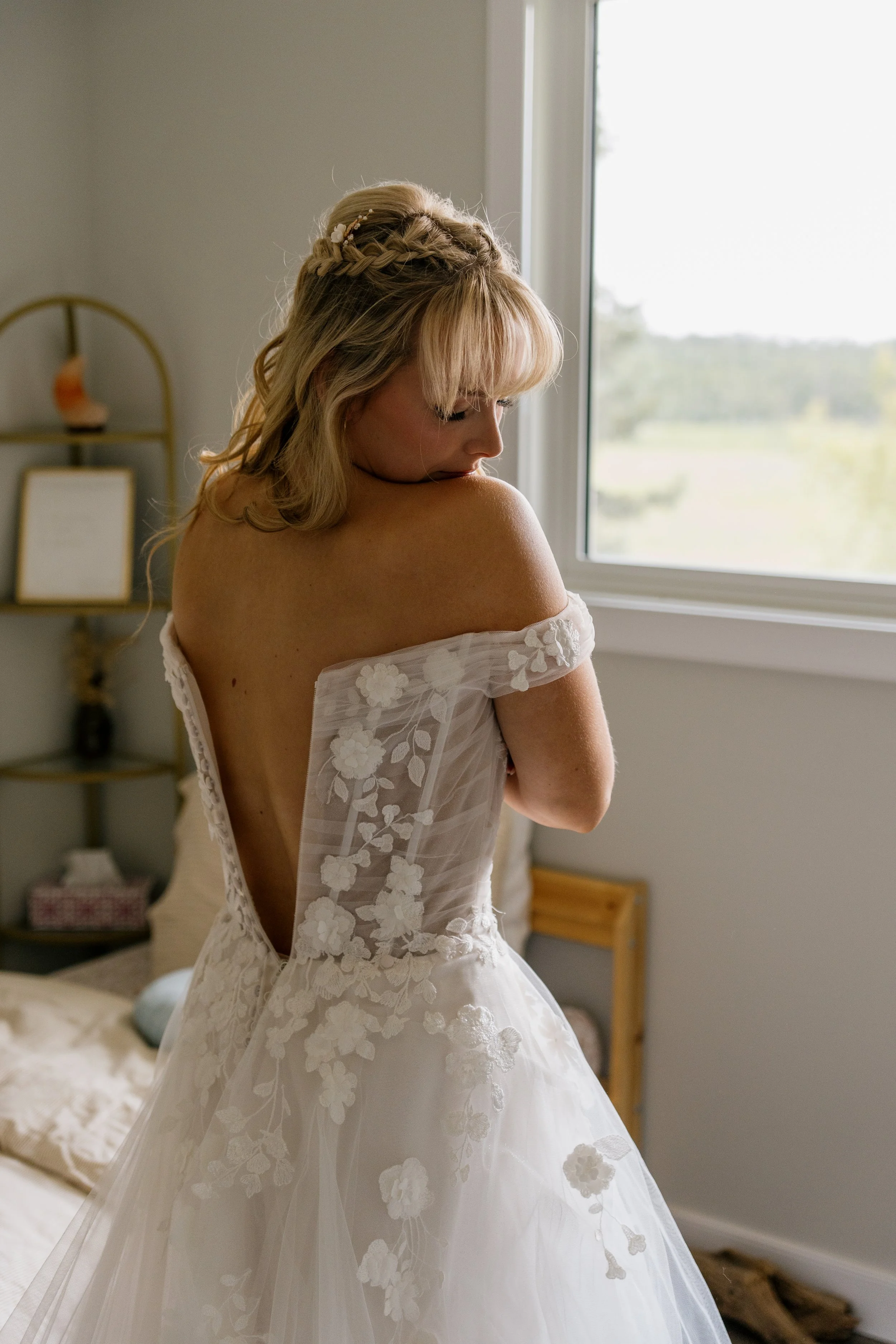 A bride in a white wedding gown with floral embroidery, standing indoors near a window, looking down with her face turned to the side.