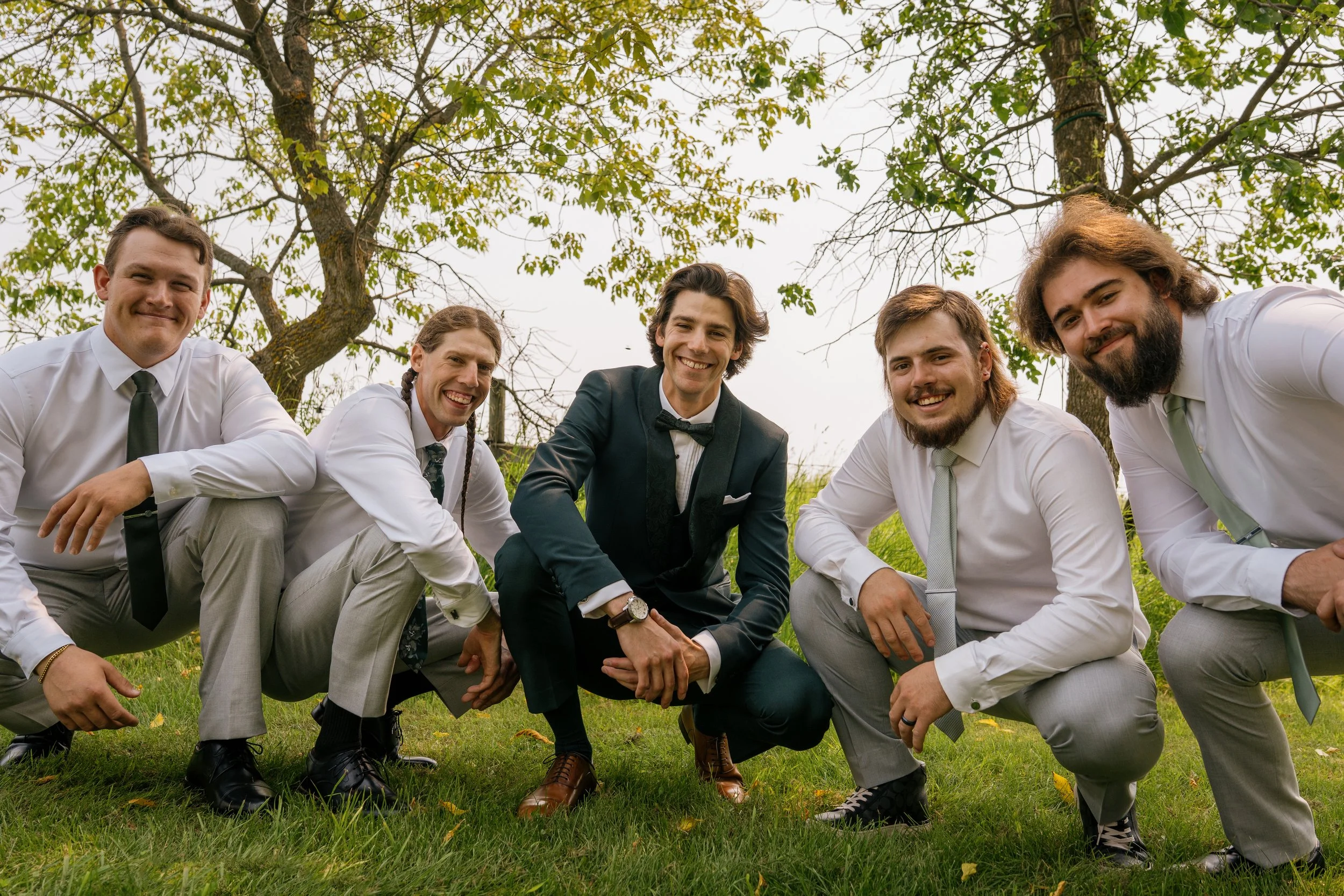 A group of six men, dressed in formal attire, are smiling and posing outdoors beneath trees with green foliage. They are crouching or kneeling on the grass.