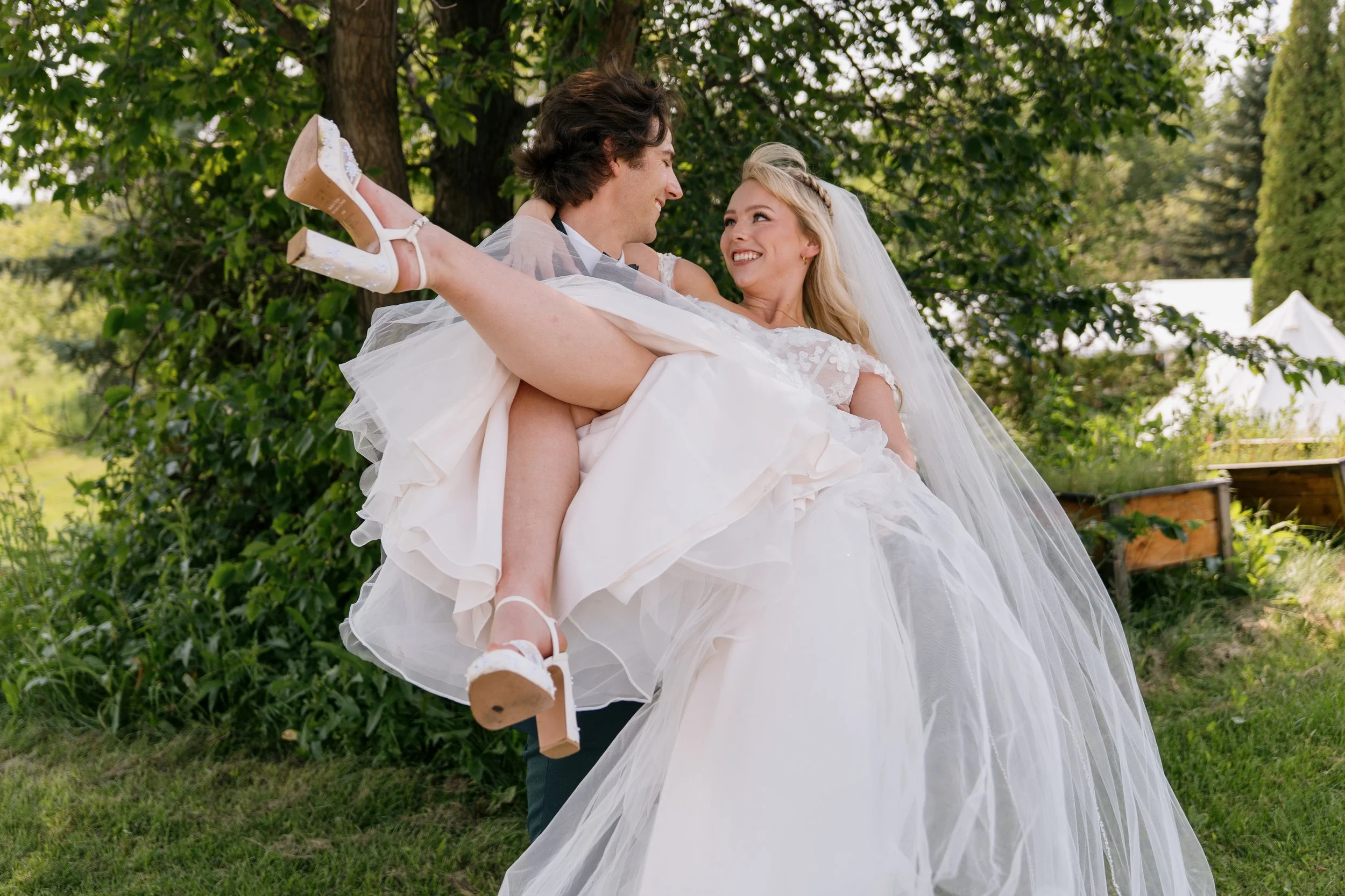 A newlywed couple outdoors; the groom in a black tuxedo carrying the smiling bride in a white wedding dress with a veil and high heels, surrounded by green trees and grass.