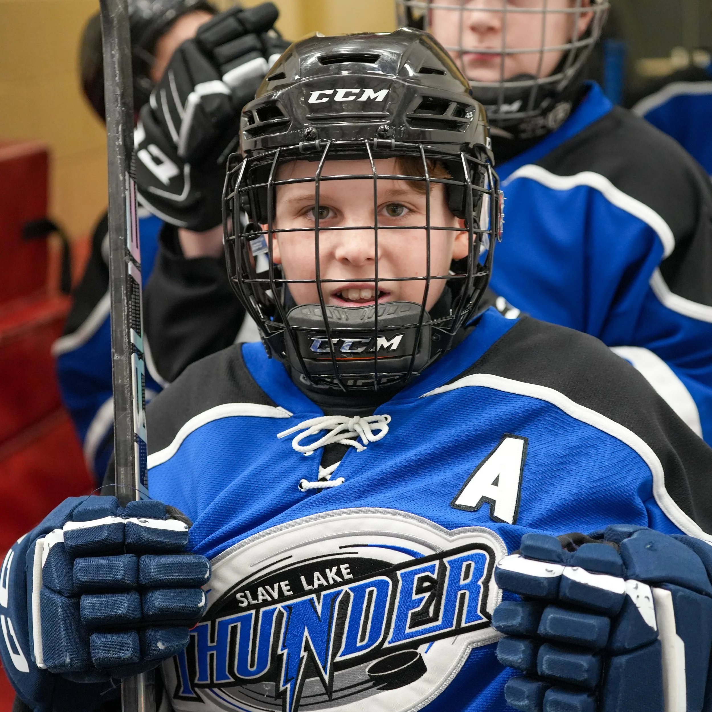 Close-up of a young hockey player in a blue jersey and black helmet, holding a hockey stick, with another player in the background wearing protective gear.