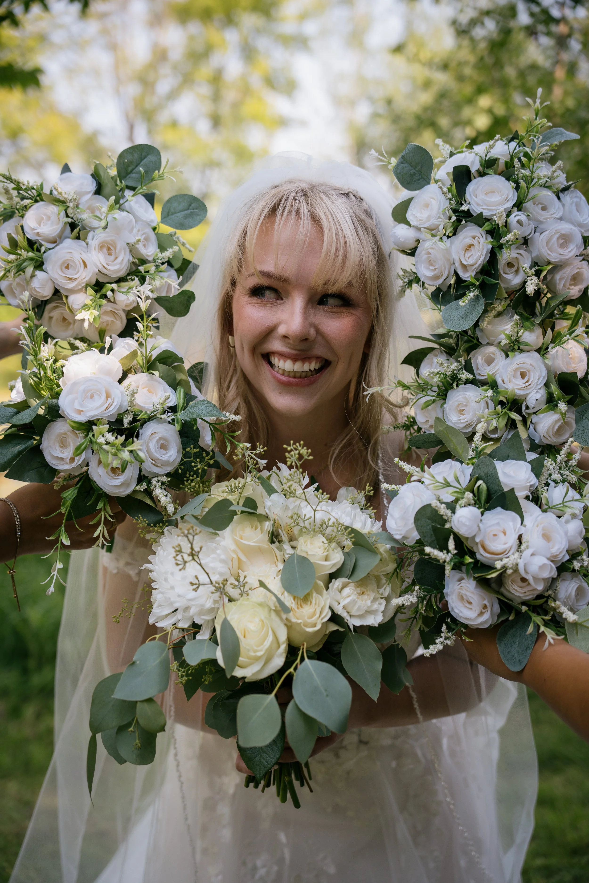 A smiling bride holding a large bouquet of white roses, greenery, and floral accents, outdoors with trees and sunlight in the background.