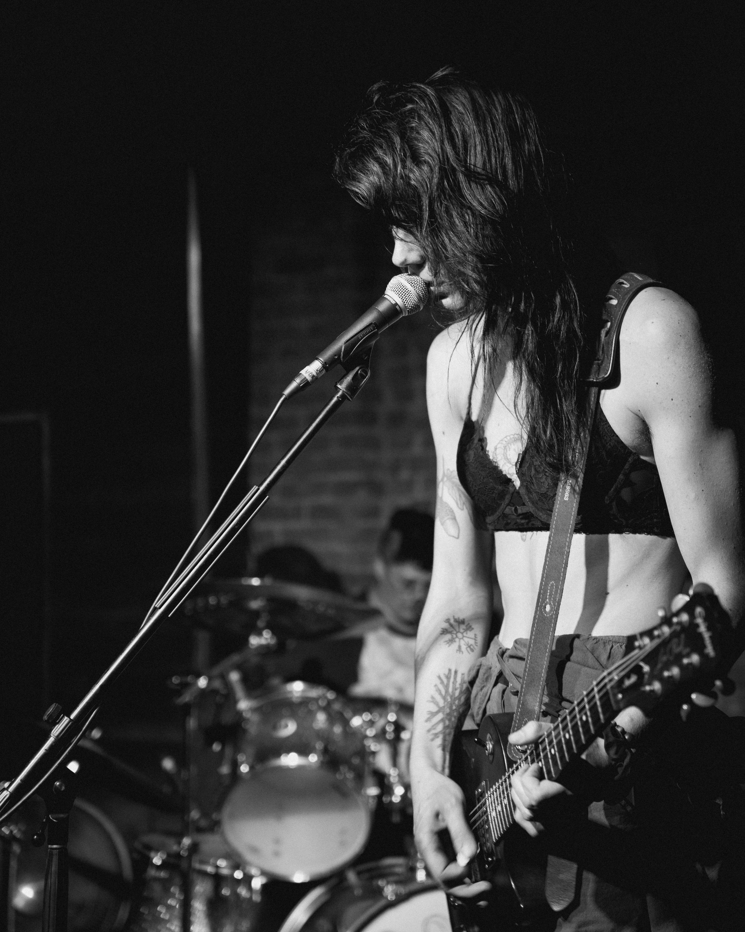 A black and white photo of a woman singing into a microphone with a guitar, with a drum set in the background.