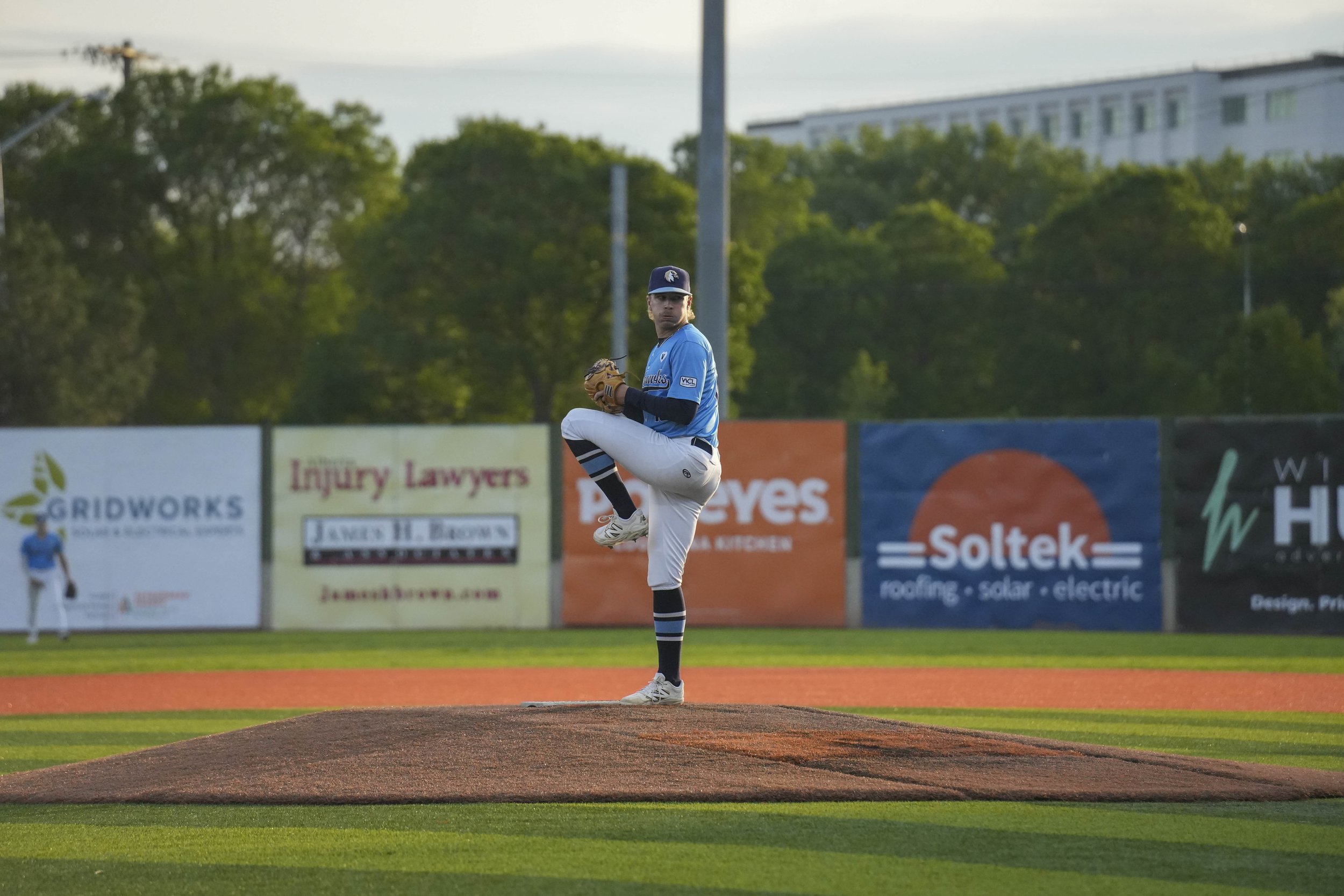 A baseball pitcher is on the mound, preparing to throw a pitch during a game, with a field and advertisements visible in the background.