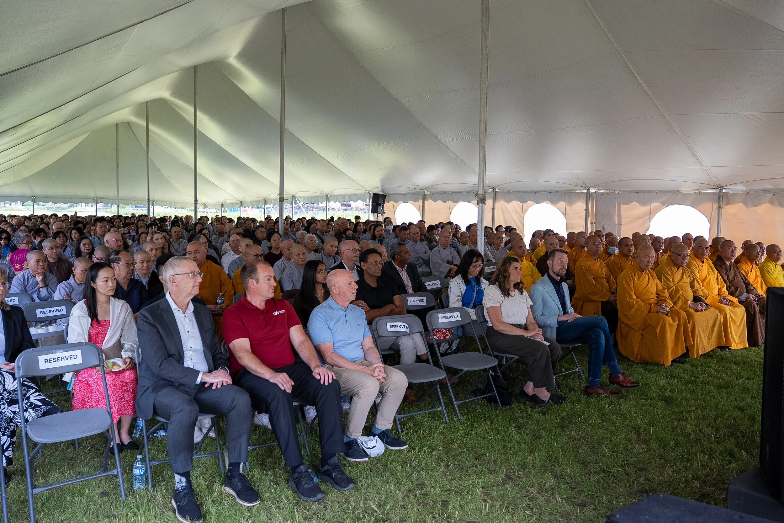 A large group of people seated under a spacious outdoor tent, attending an event, with some wearing traditional clothing and many with shaved heads, suggesting a cultural or religious gathering.
