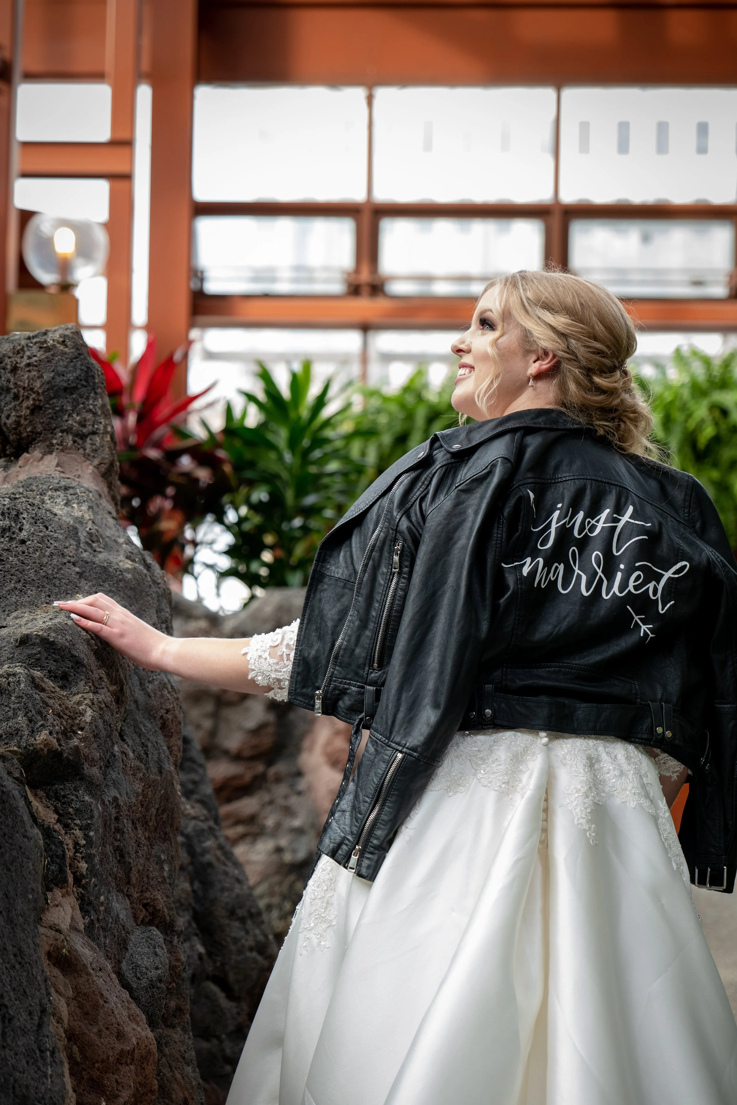 A bride wearing a white wedding dress with lace details and a black leather jacket with the words 'just married' embroidered on the back. She is touching a large rock with her left hand and smiling, with greenery and a wooden structure in the backgro