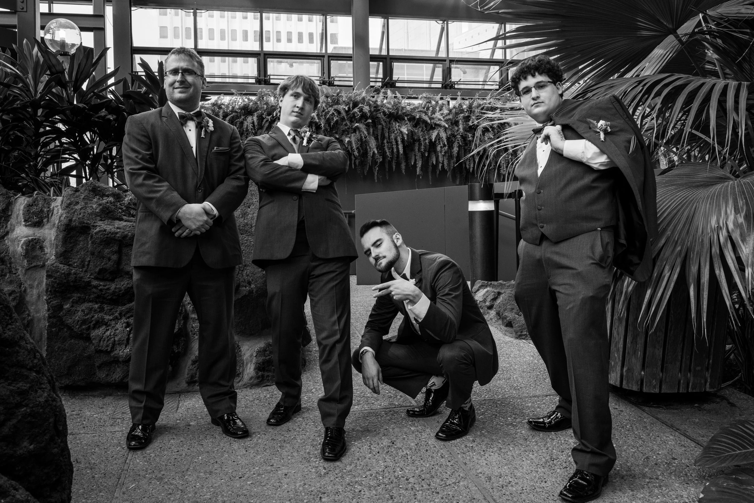 Five young men in tuxedos posing inside a modern building with plants and large windows.