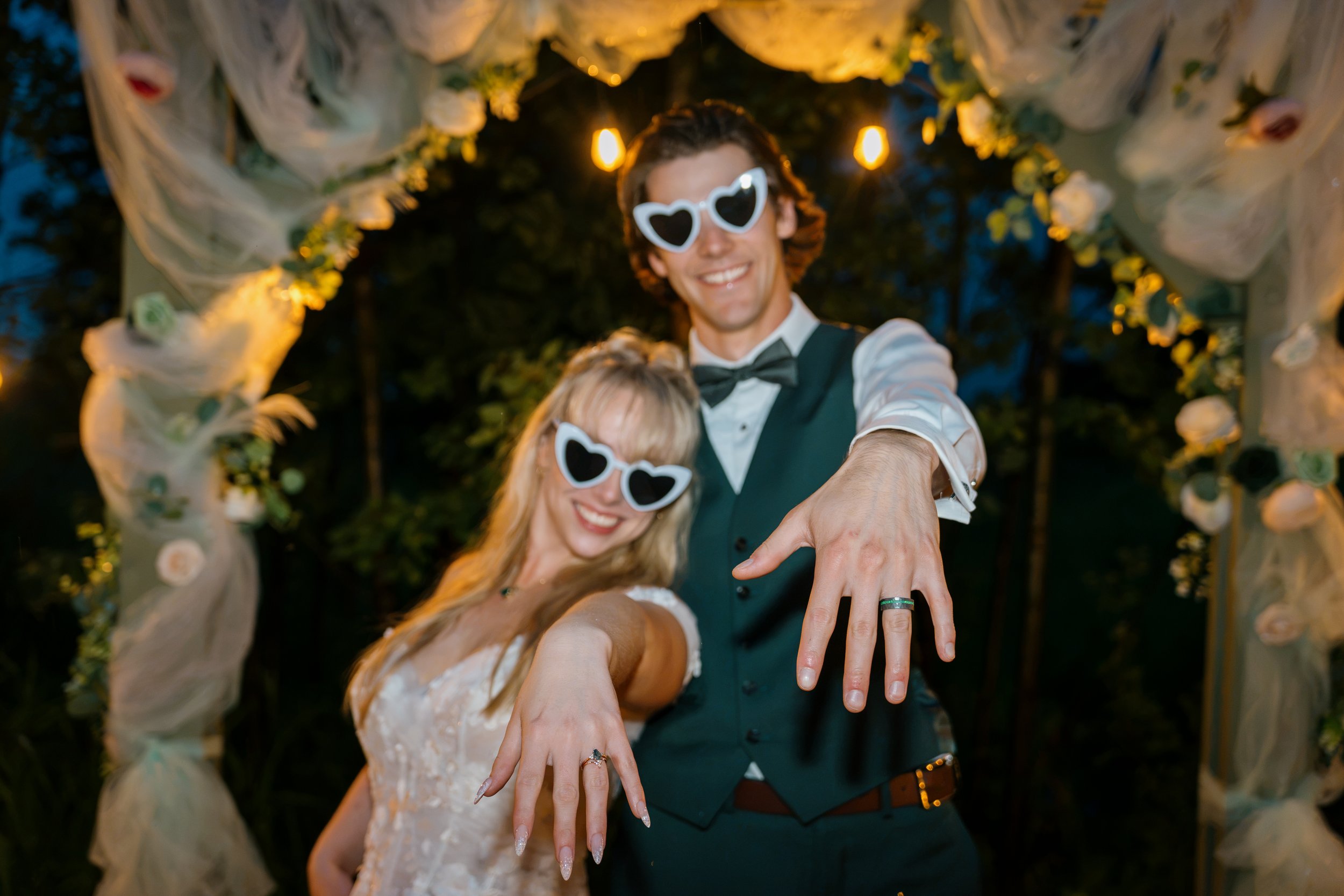 A smiling couple wearing matching heart-shaped sunglasses at a wedding or celebration, with a decorated archway in the background.