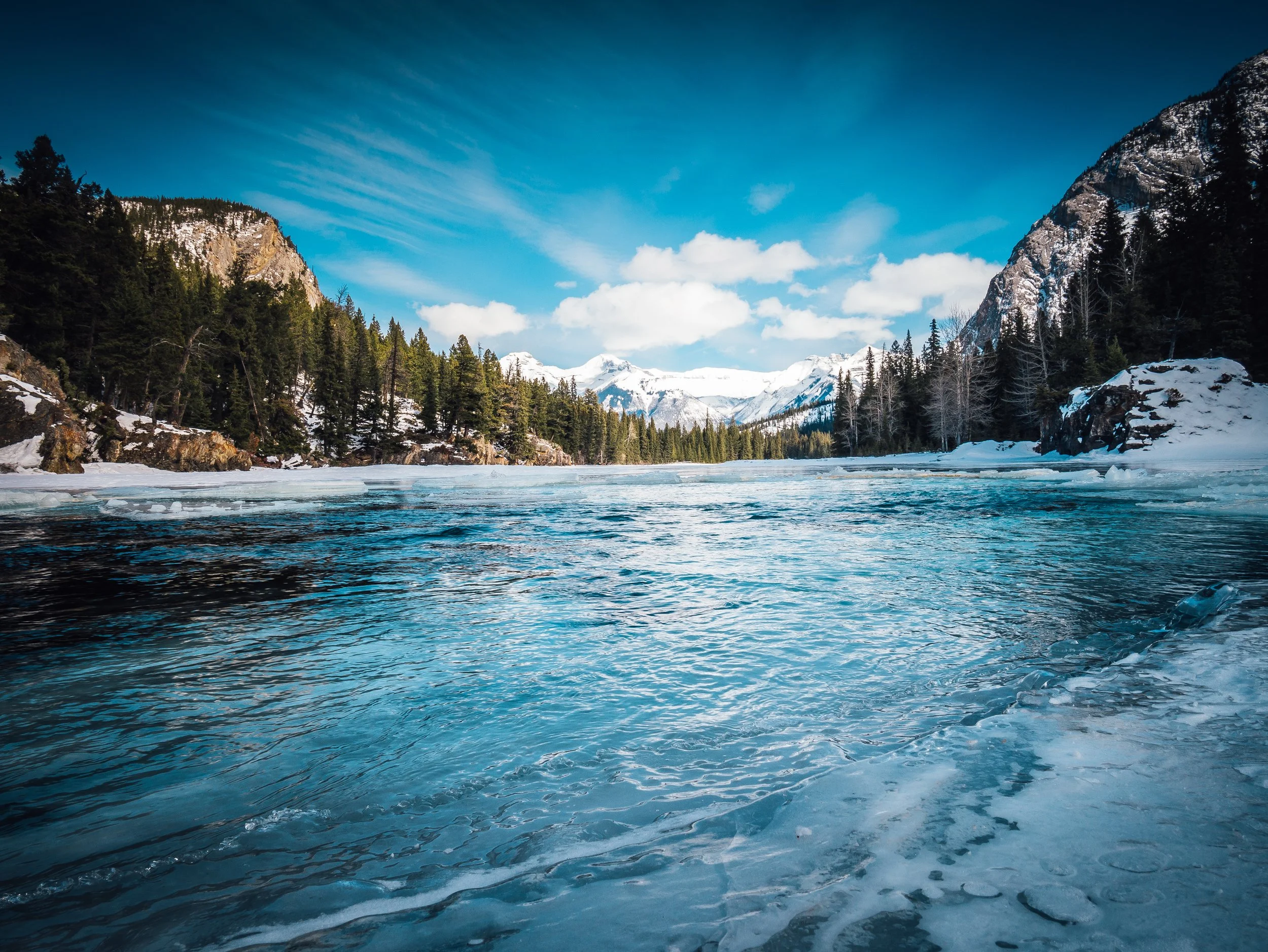 Snow-covered mountains and pine trees surrounding a partially frozen river under a blue sky with clouds.