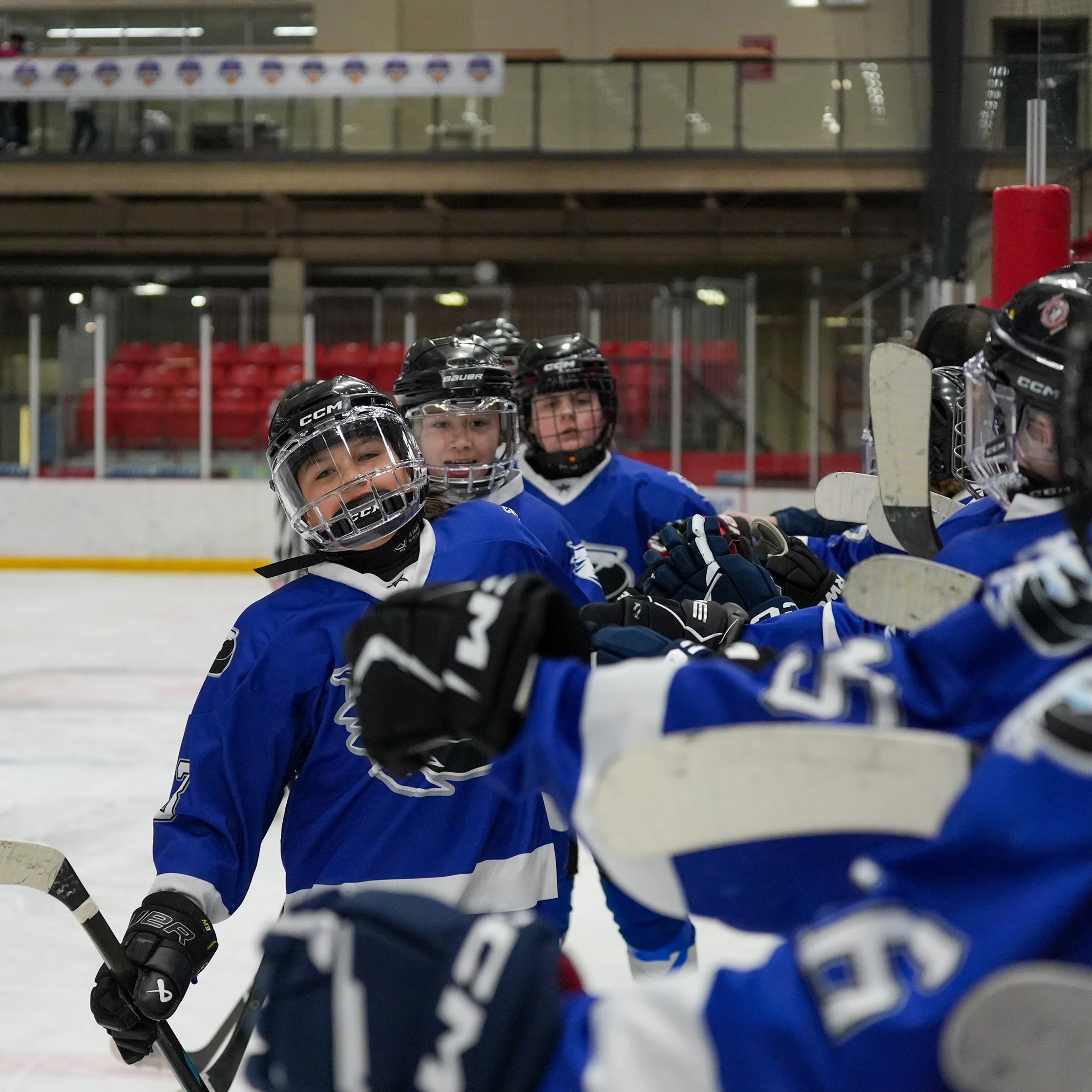 A group of young boys in hockey gear, including helmets and jerseys, sitting on the ice at an indoor hockey rink.