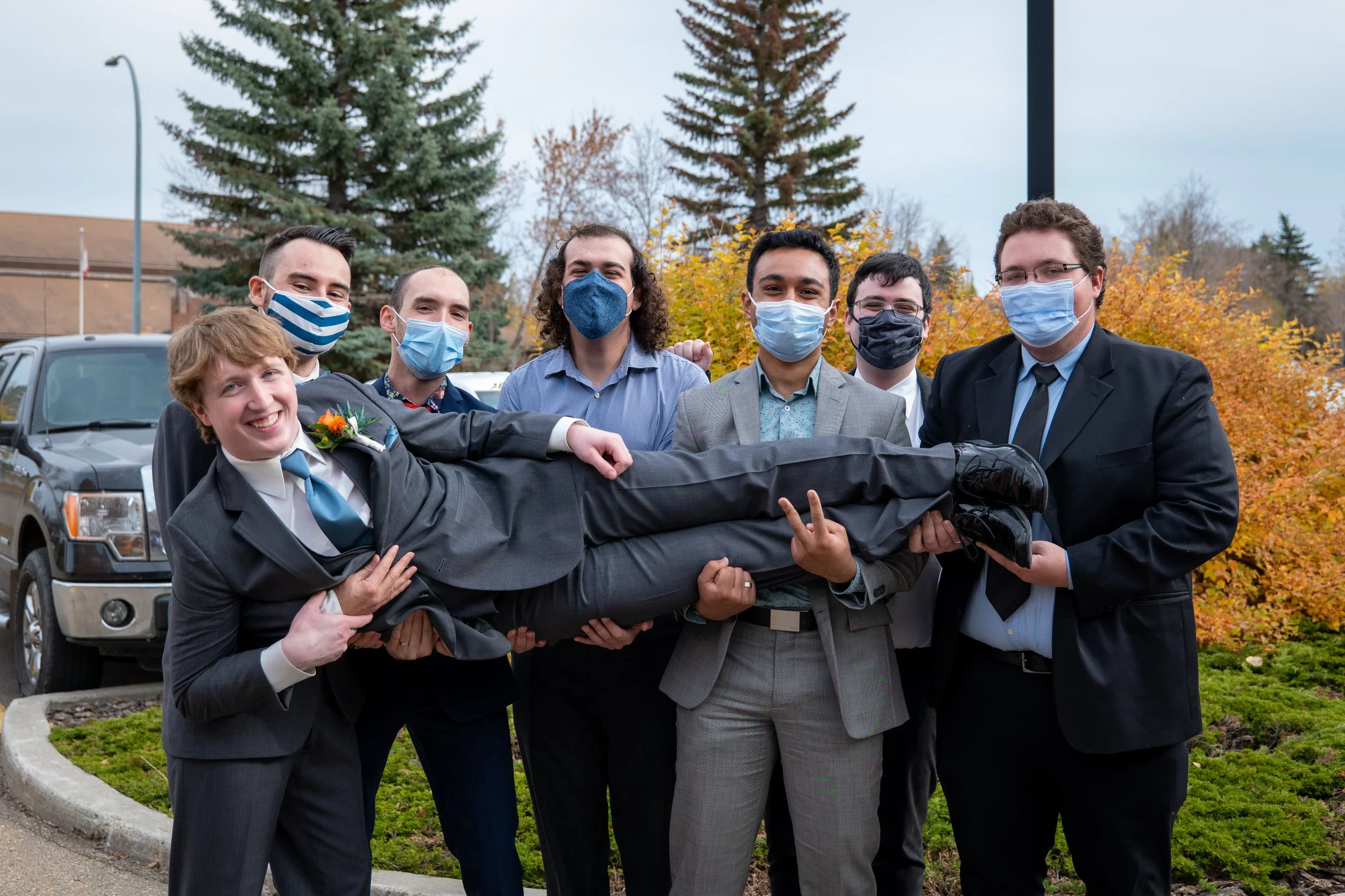 A group of seven young men in suits and masks are standing outdoors in front of trees with fall foliage. They are holding a man in a suit horizontally, who is smiling and making a peace sign with his hand.