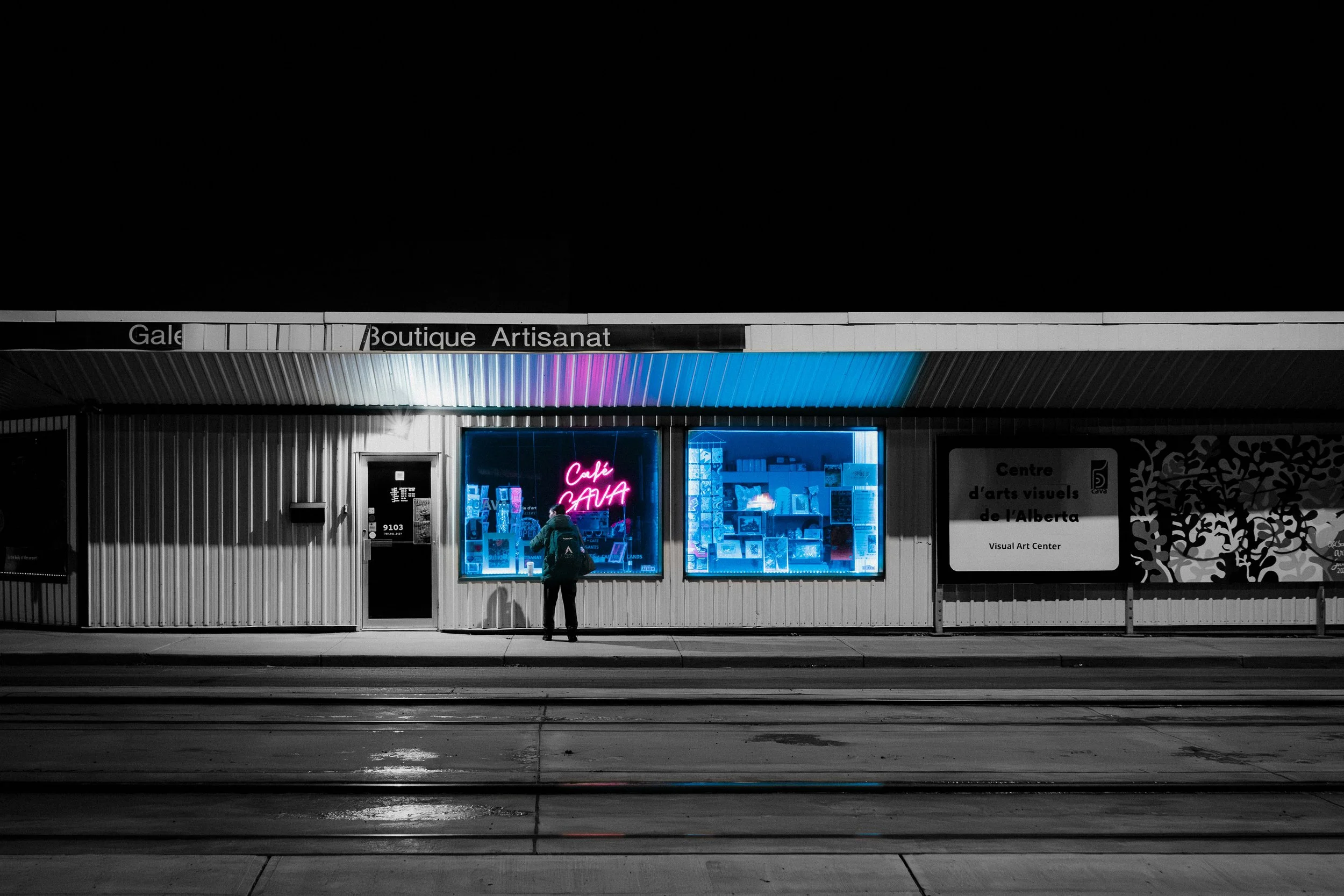 Nighttime view of a storefront with neon signs, one reading 'Café CAVA,' and another advertising an arts center, with a person standing outside.