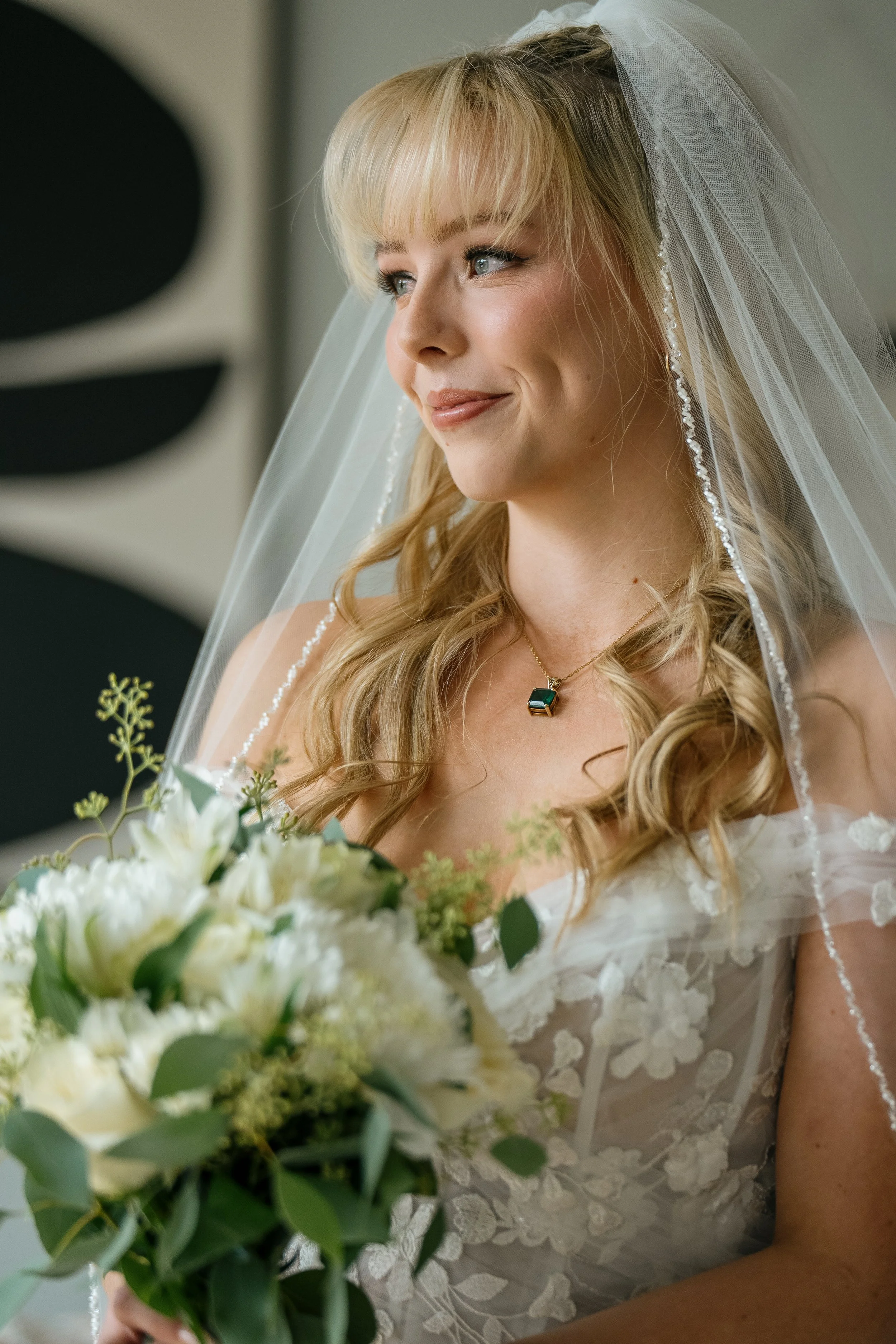 A bride with blonde hair in loose curls, wearing a white lace wedding dress and a veil, holding a bouquet of white flowers and greenery, standing indoors.