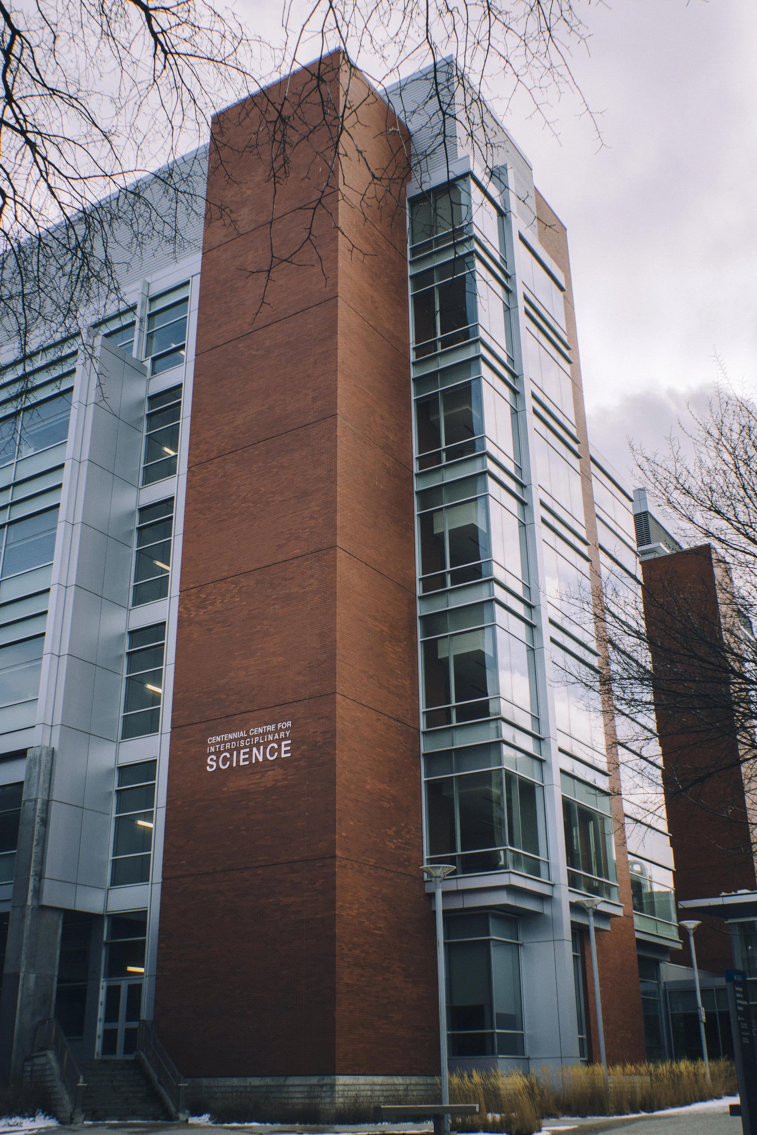 Tall modern building made of brick and glass with the sign 'Centennial Centre for Interdisciplinary Science' near the bottom, under a cloudy sky, surrounded by leafless trees and outdoor light poles.