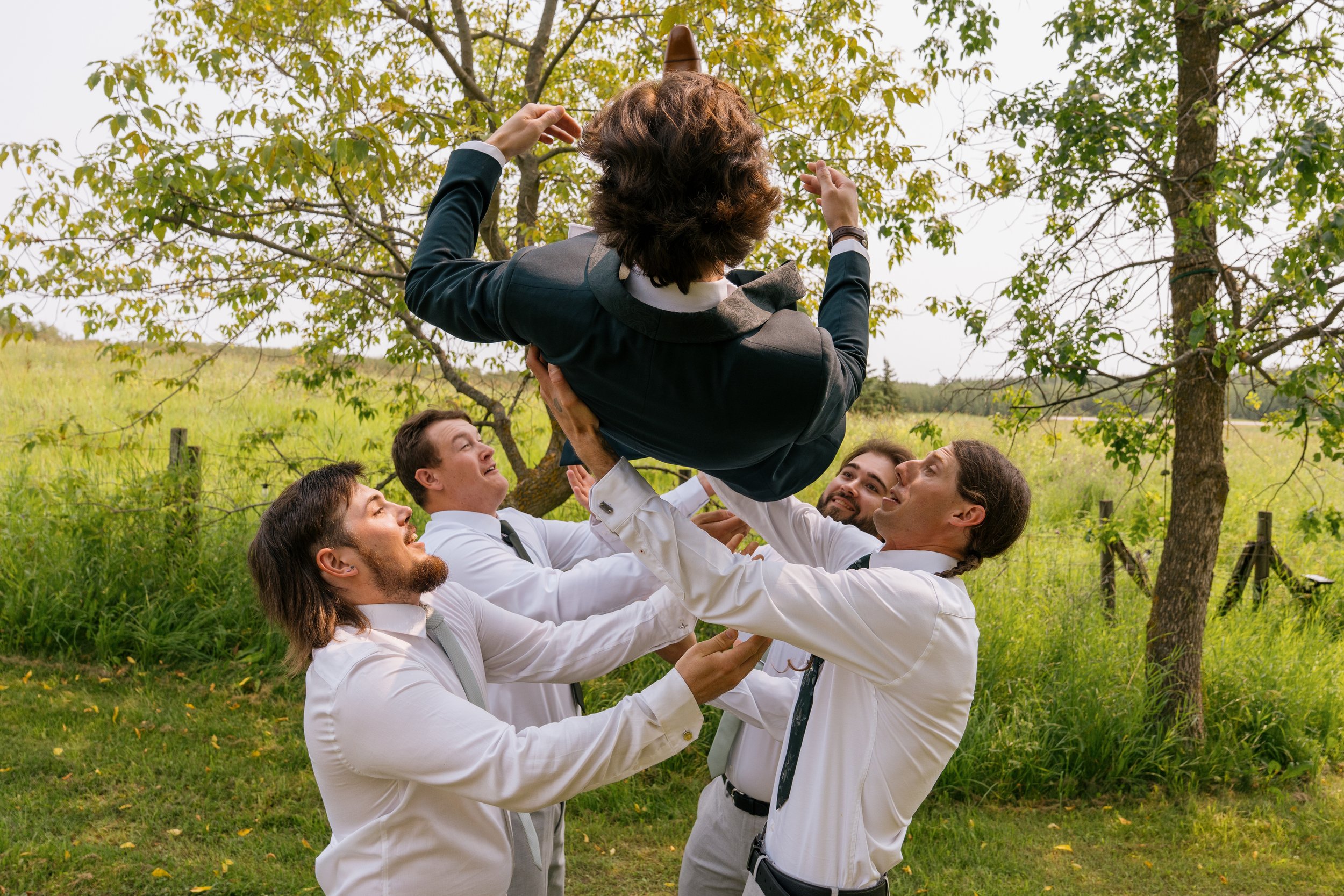 A group of five men in white shirts and ties lifting a woman in a suit and skirt with a brown hat in the air outdoors among trees and green grass.
