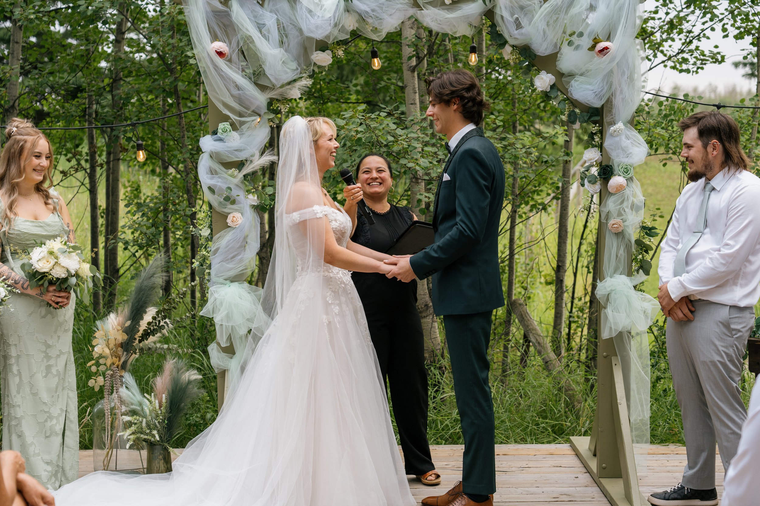A wedding ceremony taking place outdoors among trees with a couple holding hands and smiling at each other. The bride is wearing a white wedding dress and veil, and the groom is dressed in a dark suit. An officiant is standing between them, holding a