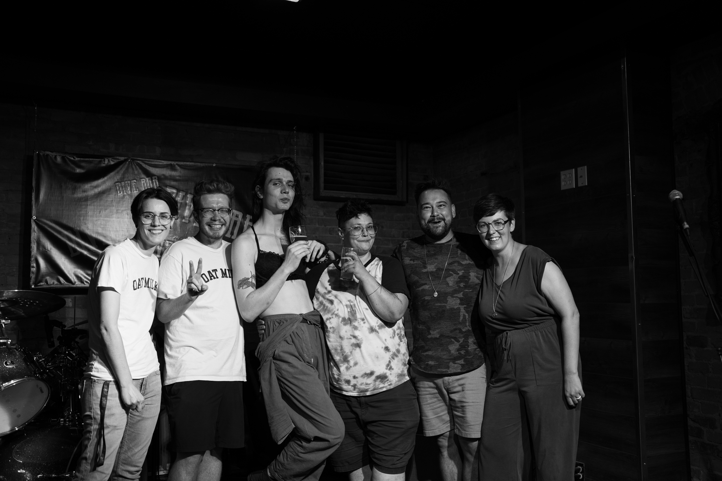 Group of six people posing together on stage, holding glasses, with a drum set in the background, in a dimly lit indoor venue.