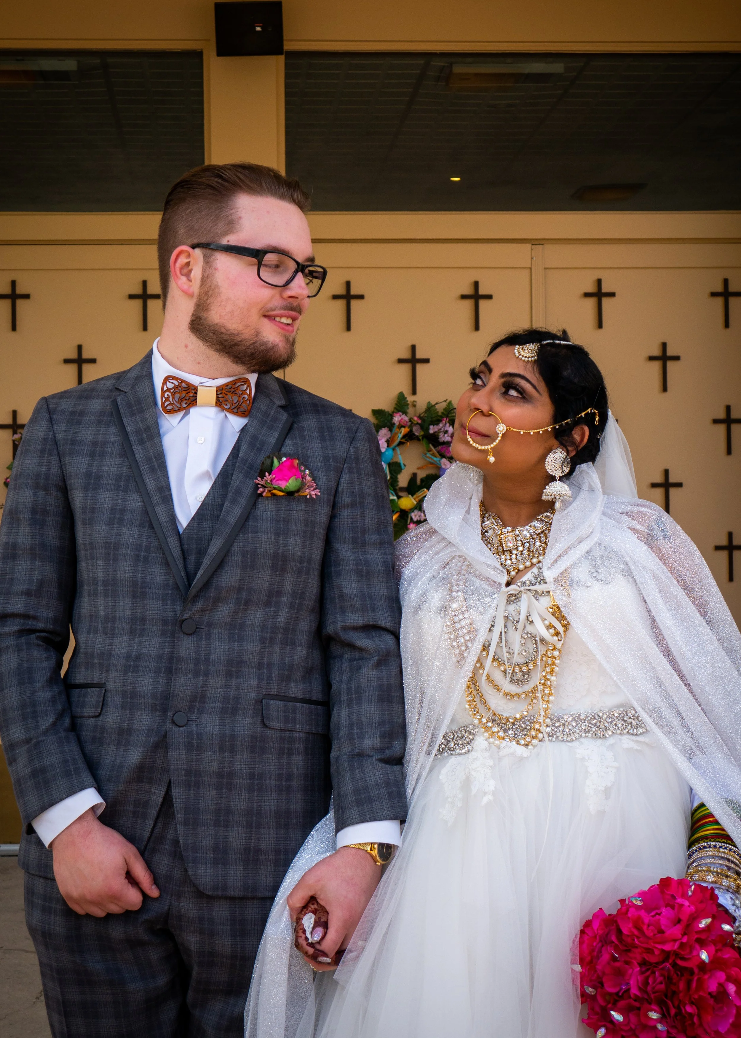 A bride and groom holding hands during their wedding ceremony, standing in front of a cross-decorated wall indoors.