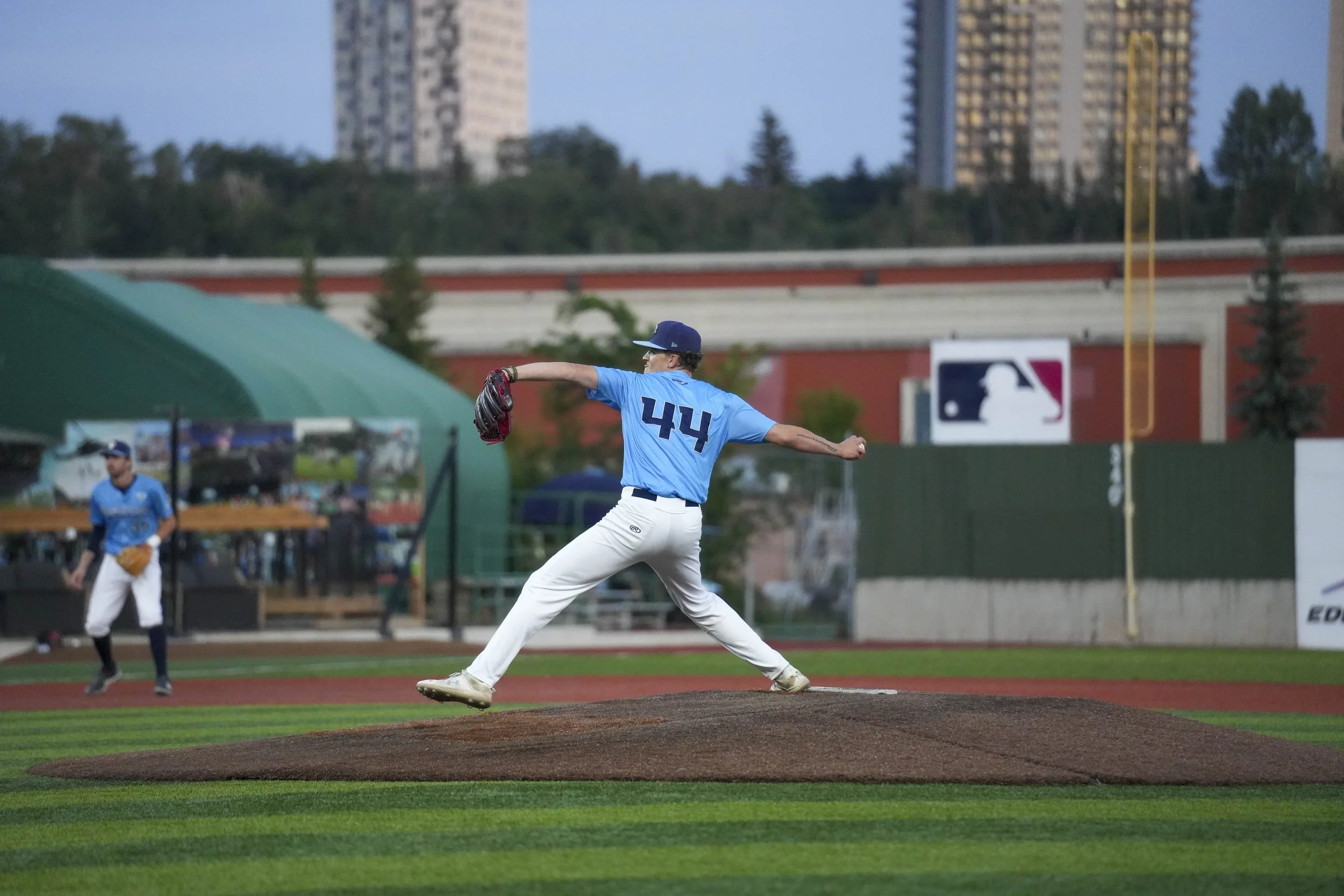 A baseball player in a blue jersey with number 44 is pitching on the mound at a baseball field, with a teammate in the background and city buildings beyond the field during evening.