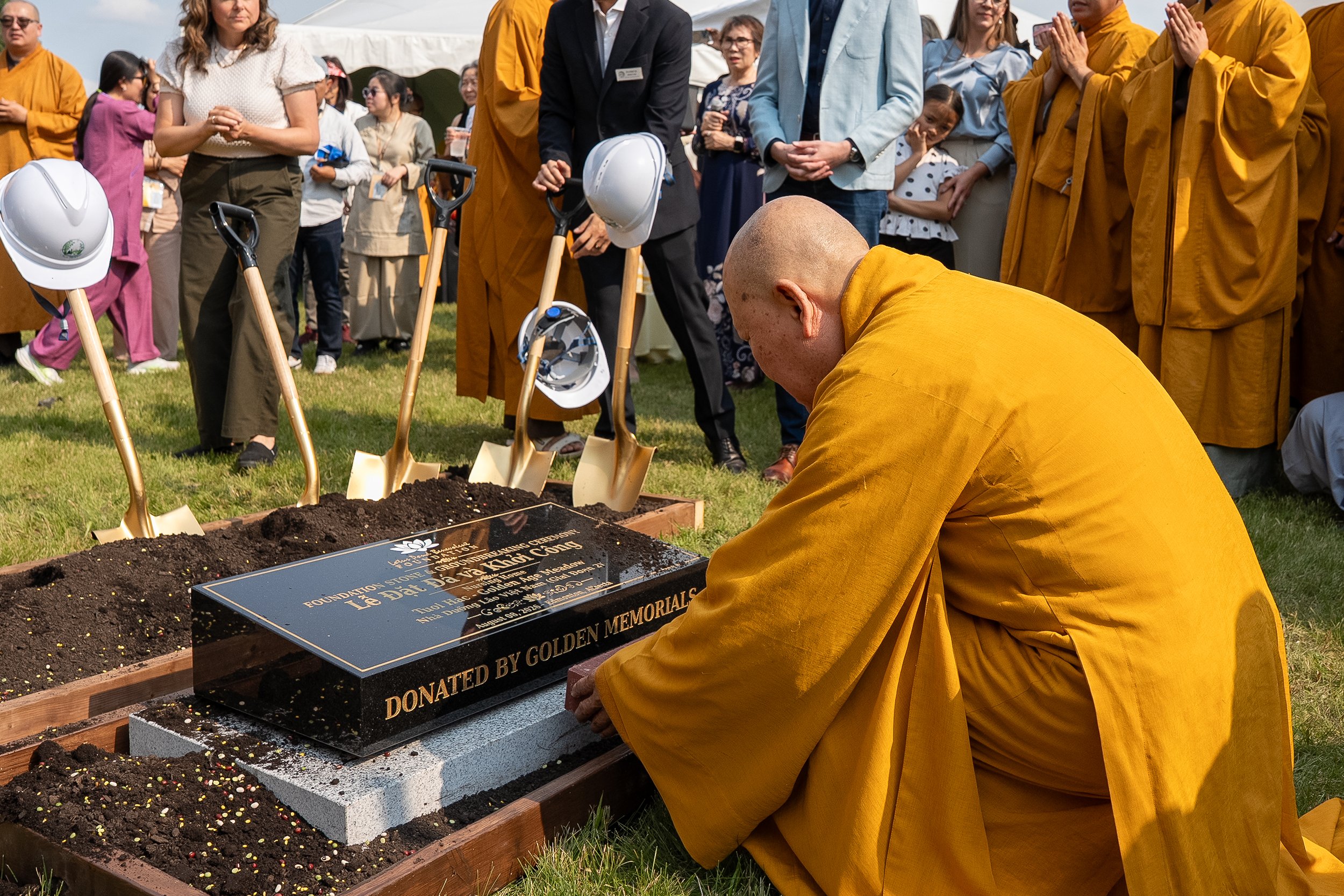 A Buddhist monk in yellow robes kneels at a memorial plaque during a memorial ceremony attended by a diverse group of people, including monks, women, and children, outdoors on a grassy area with a blue sky.