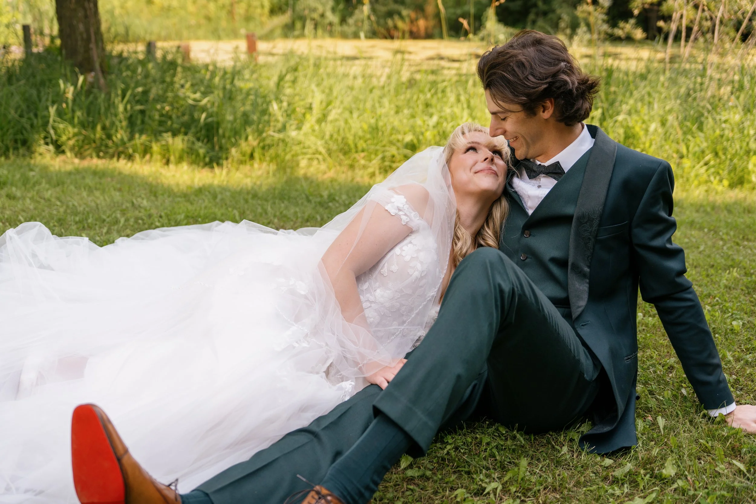 A bride and groom sit on grass outdoors. The bride rests her head on the groom's shoulder as they smile and look at each other, with a blurred green background.