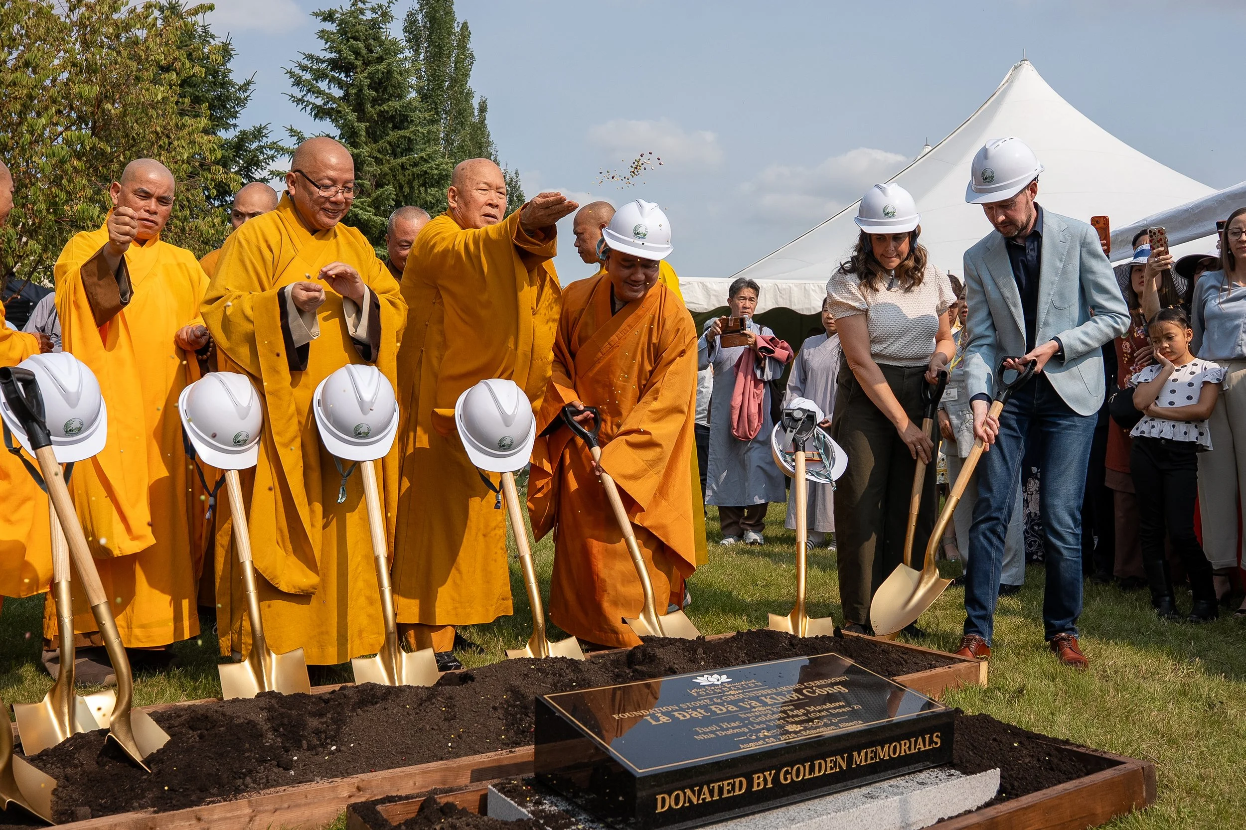 Dignitaries and monks participate in a groundbreaking ceremony with shovels, marking the start of a construction project at a memorial site, with a tent and trees in the background.