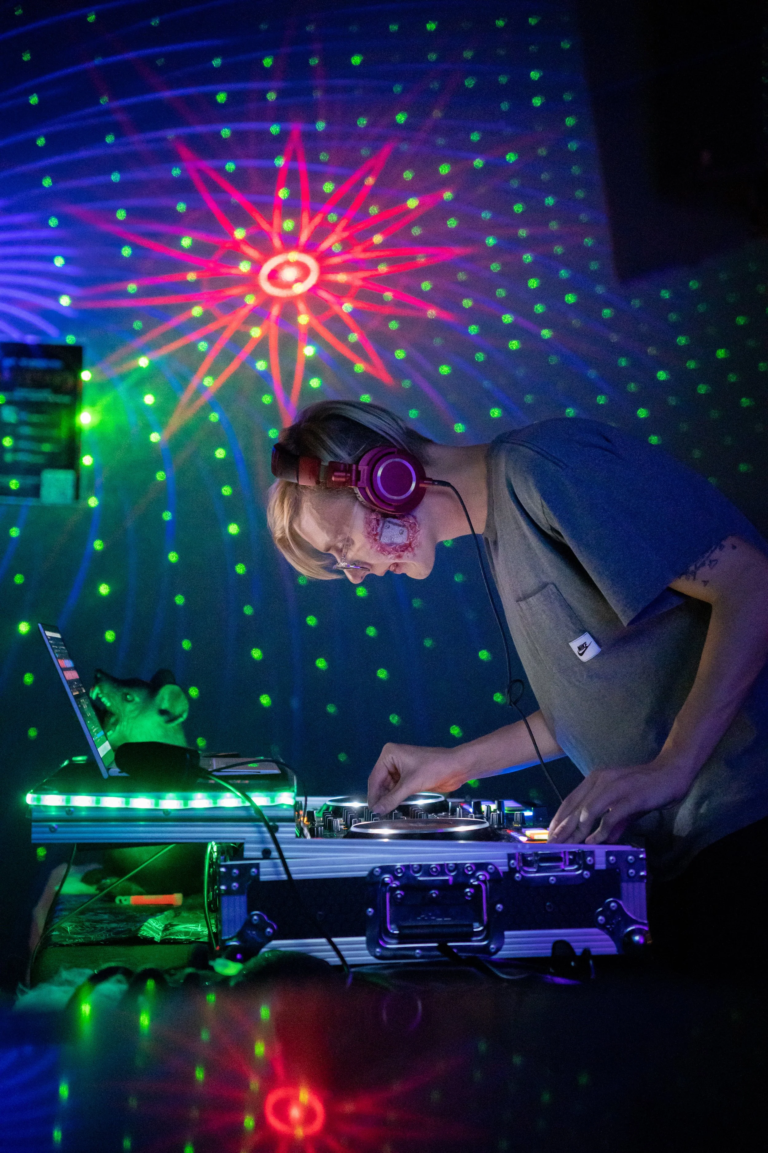 A DJ wearing headphones, mixing music on a DJ console illuminated with green and purple lights, with a background of colorful laser lights creating star and dot patterns.