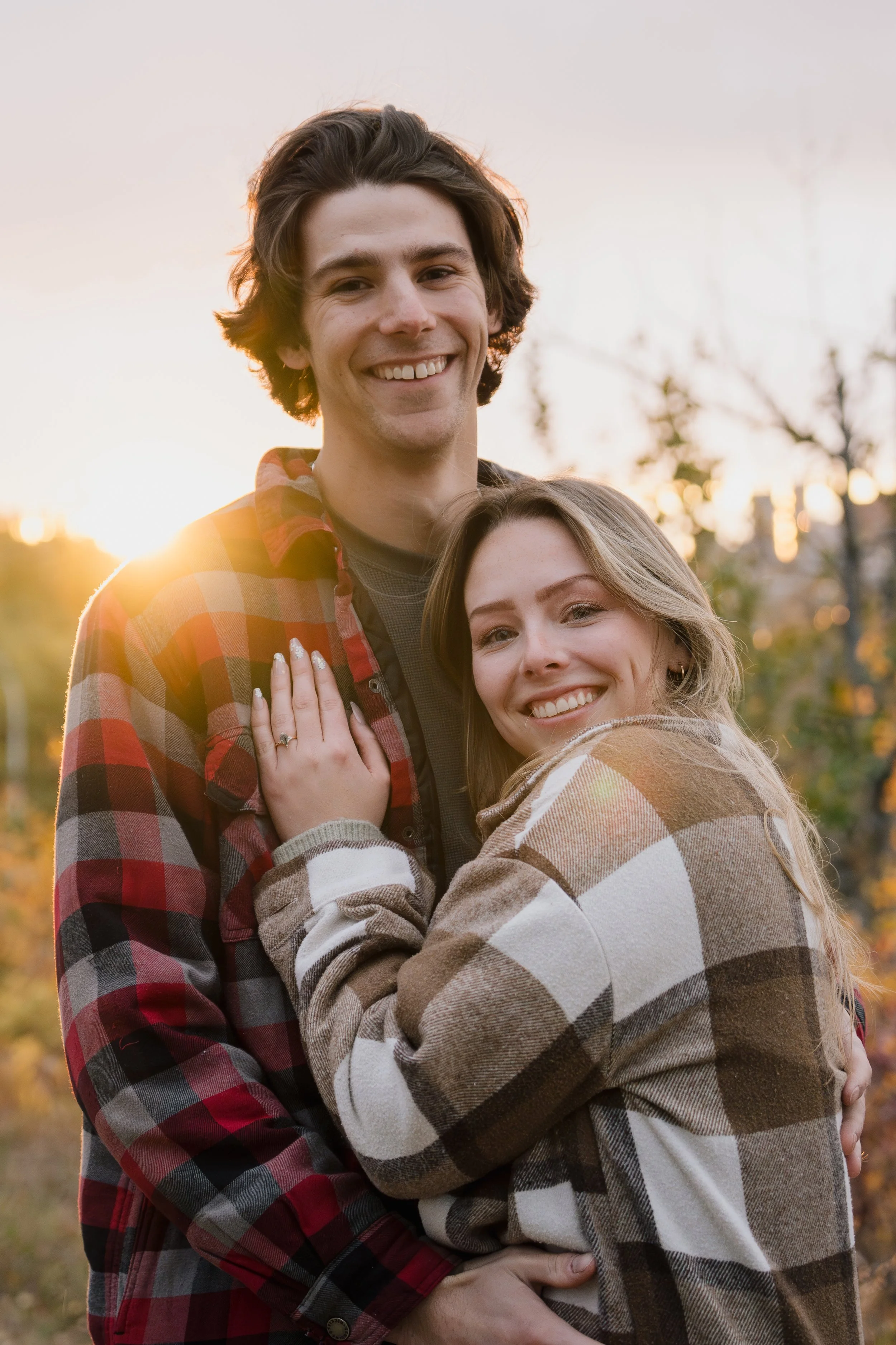 A young man and woman smiling and embracing outdoors during sunset, wearing plaid shirts.