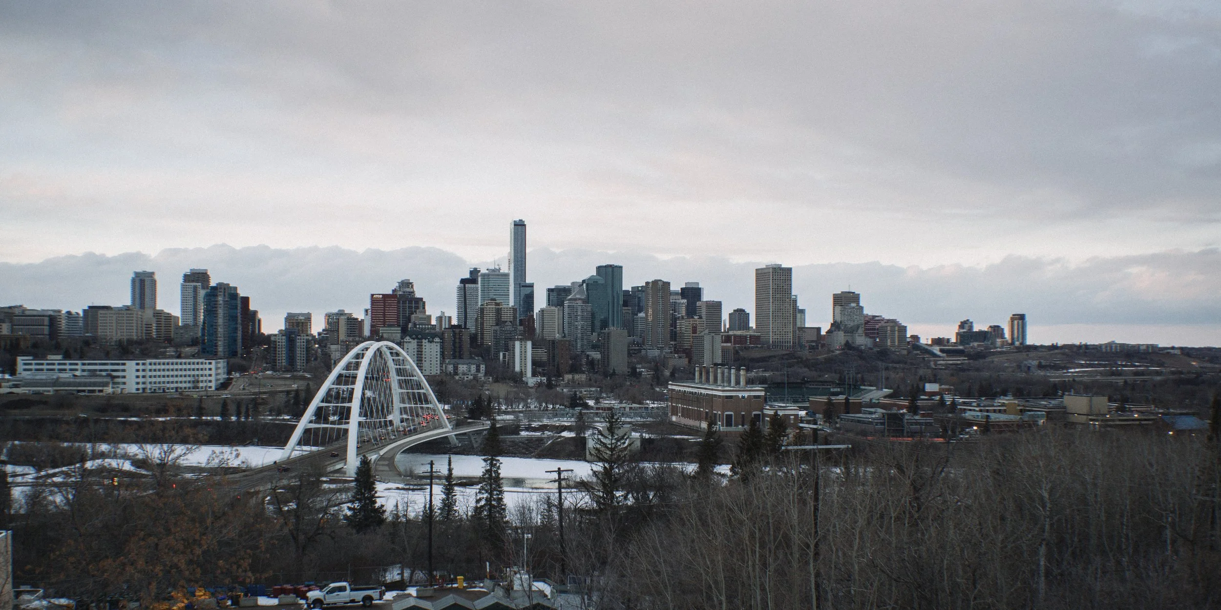 City skyline with tall buildings, a distinctive white arched bridge, and snow-covered ground under a cloudy sky.