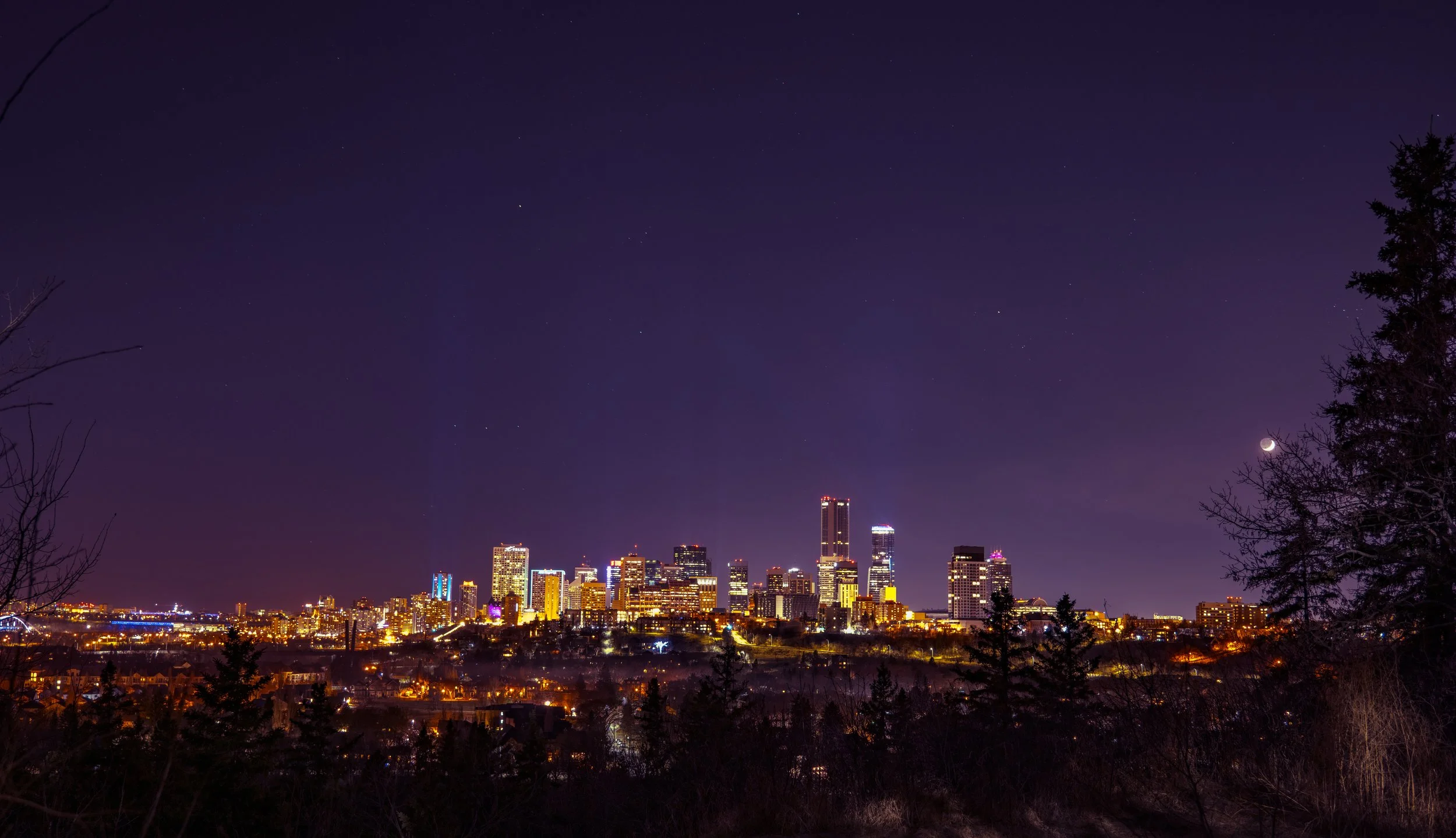 Nighttime city skyline with tall buildings illuminated, under a dark sky with stars and a crescent moon visible on the right side, framed by silhouetted trees.