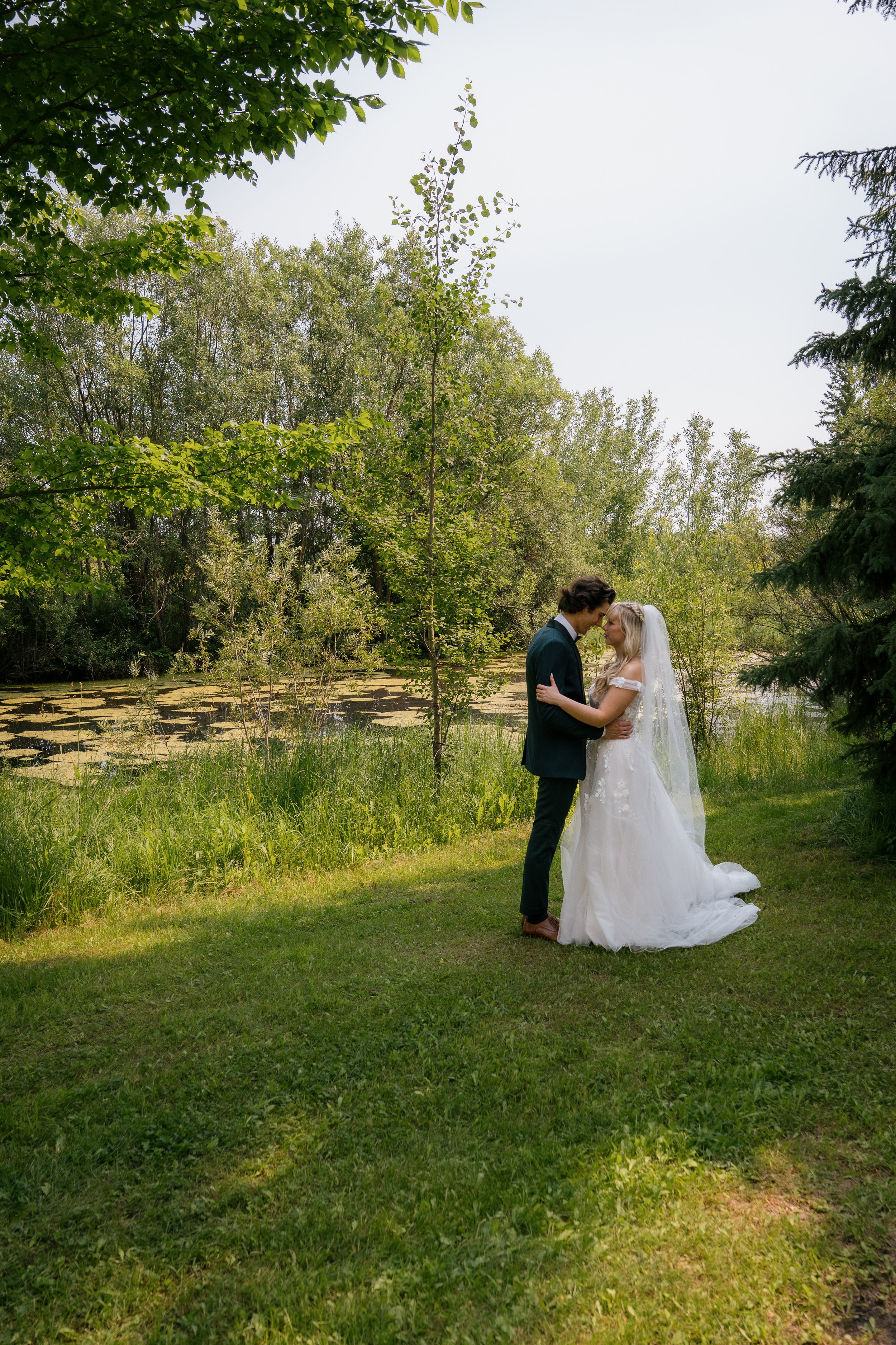 A bride and groom standing close together in a lush, green outdoor setting, holding each other and touching foreheads. The bride is wearing a white wedding gown with a long veil, and the groom is dressed in a dark suit. Behind them are trees and a po