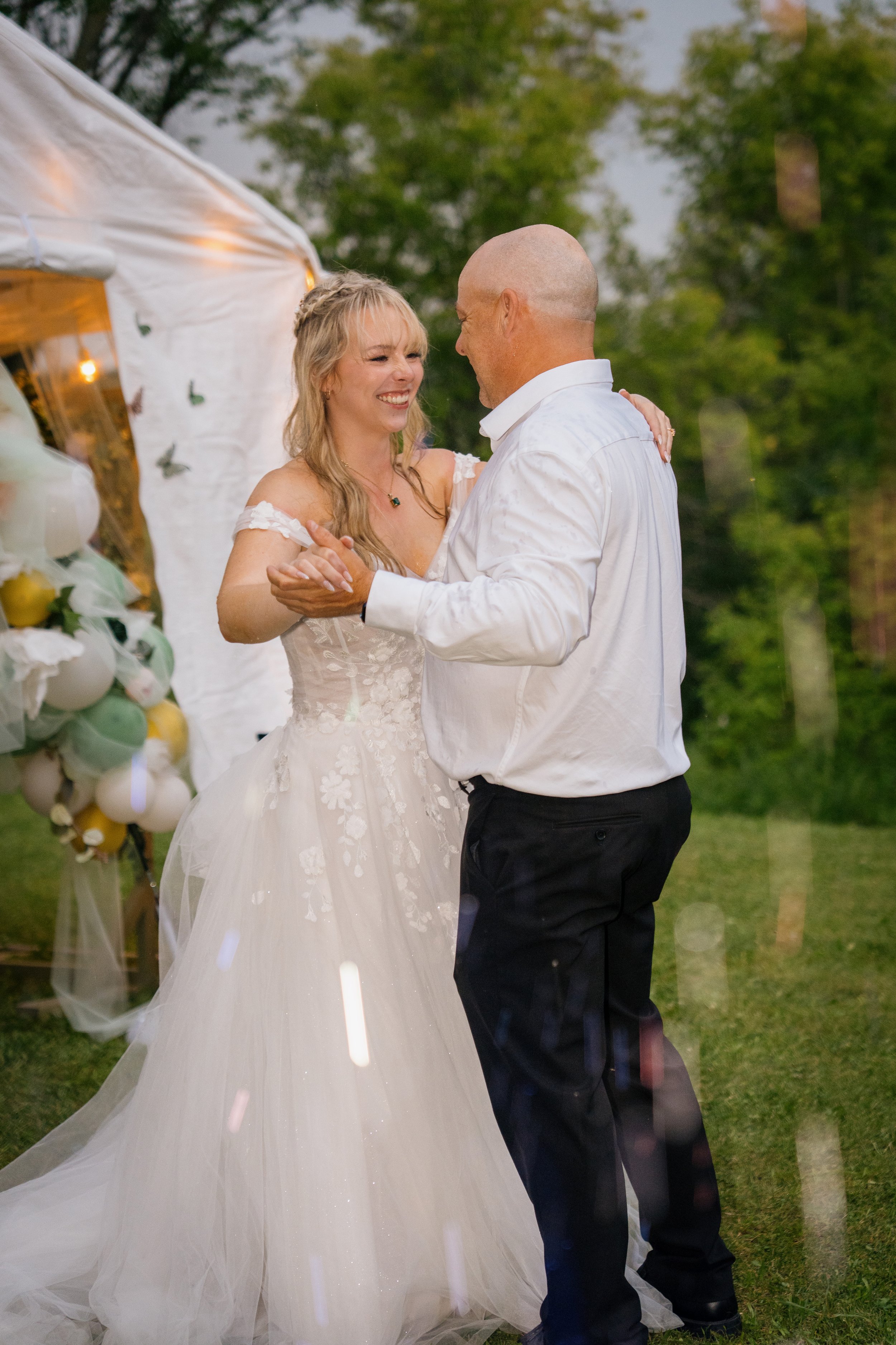 A bride and groom are dancing outdoors during their wedding, surrounded by green trees and string lights.