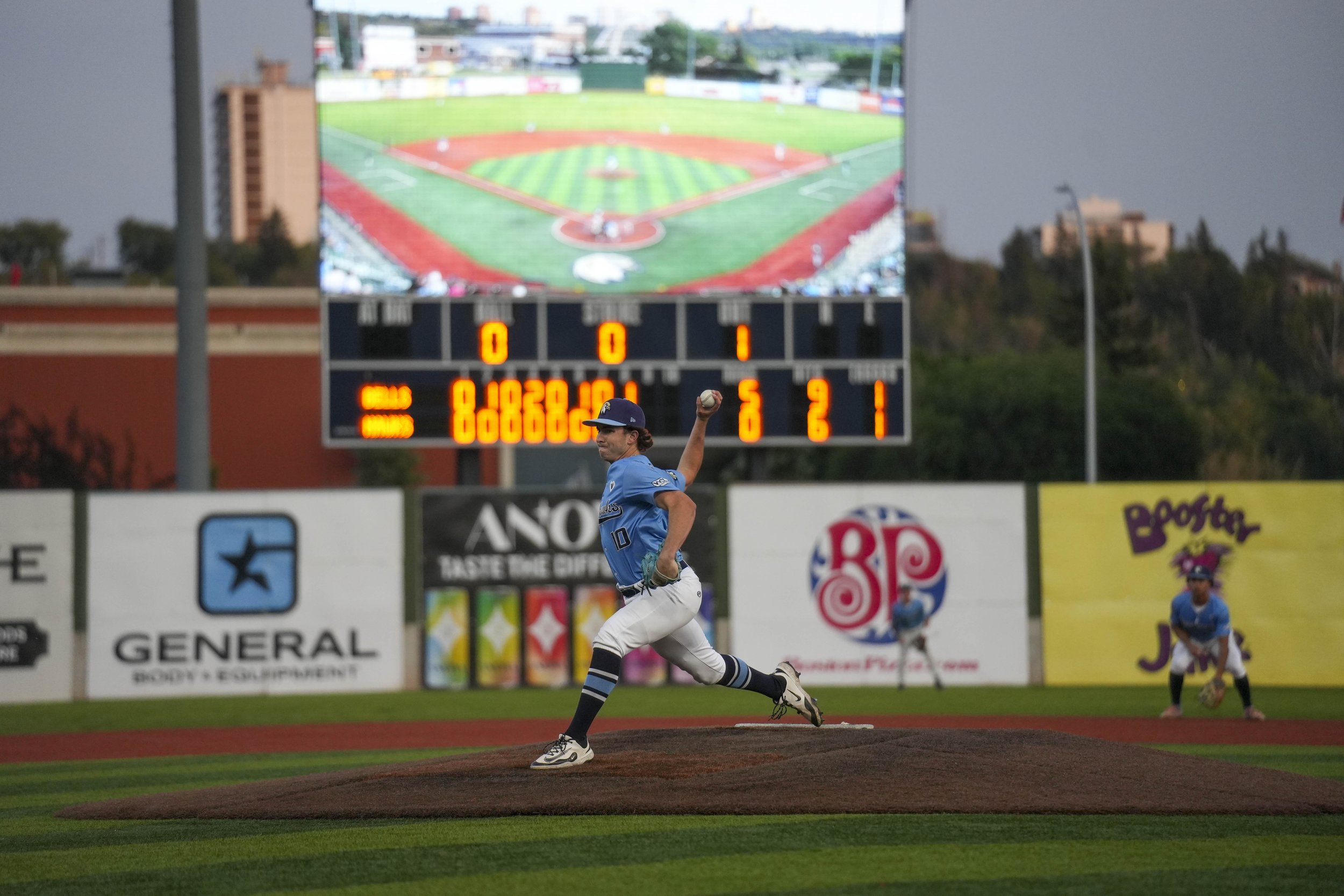 A baseball player wearing a blue jersey and cap is pitching on the mound during a game at a baseball stadium. There is a large digital scoreboard in the background showing the game score and statistics. The stadium is open with a scenic view of trees