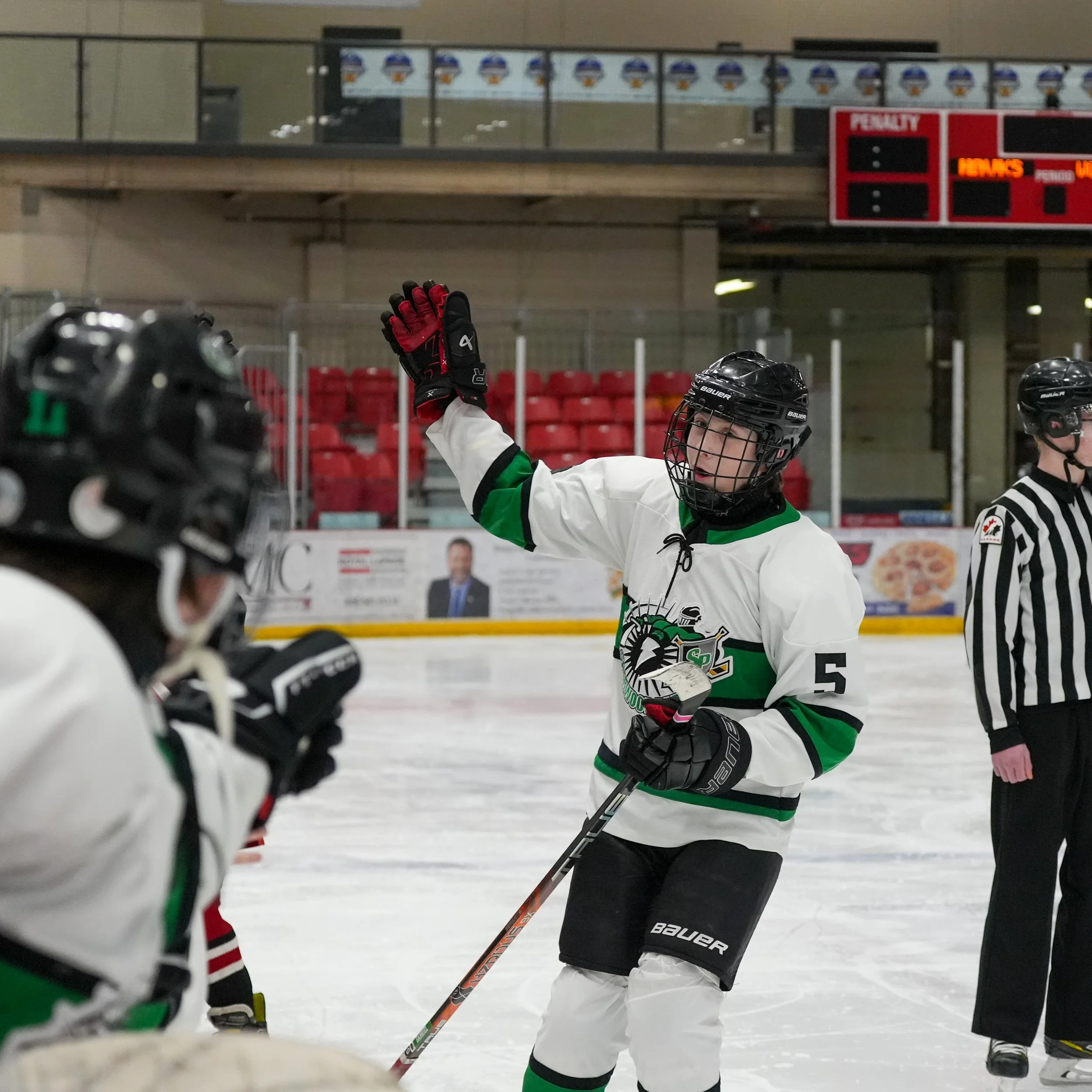 Ice hockey players on the rink, one raising a gloved hand in celebration. Player is wearing a white jersey with green and black accents, a black helmet, and holding a hockey stick.
