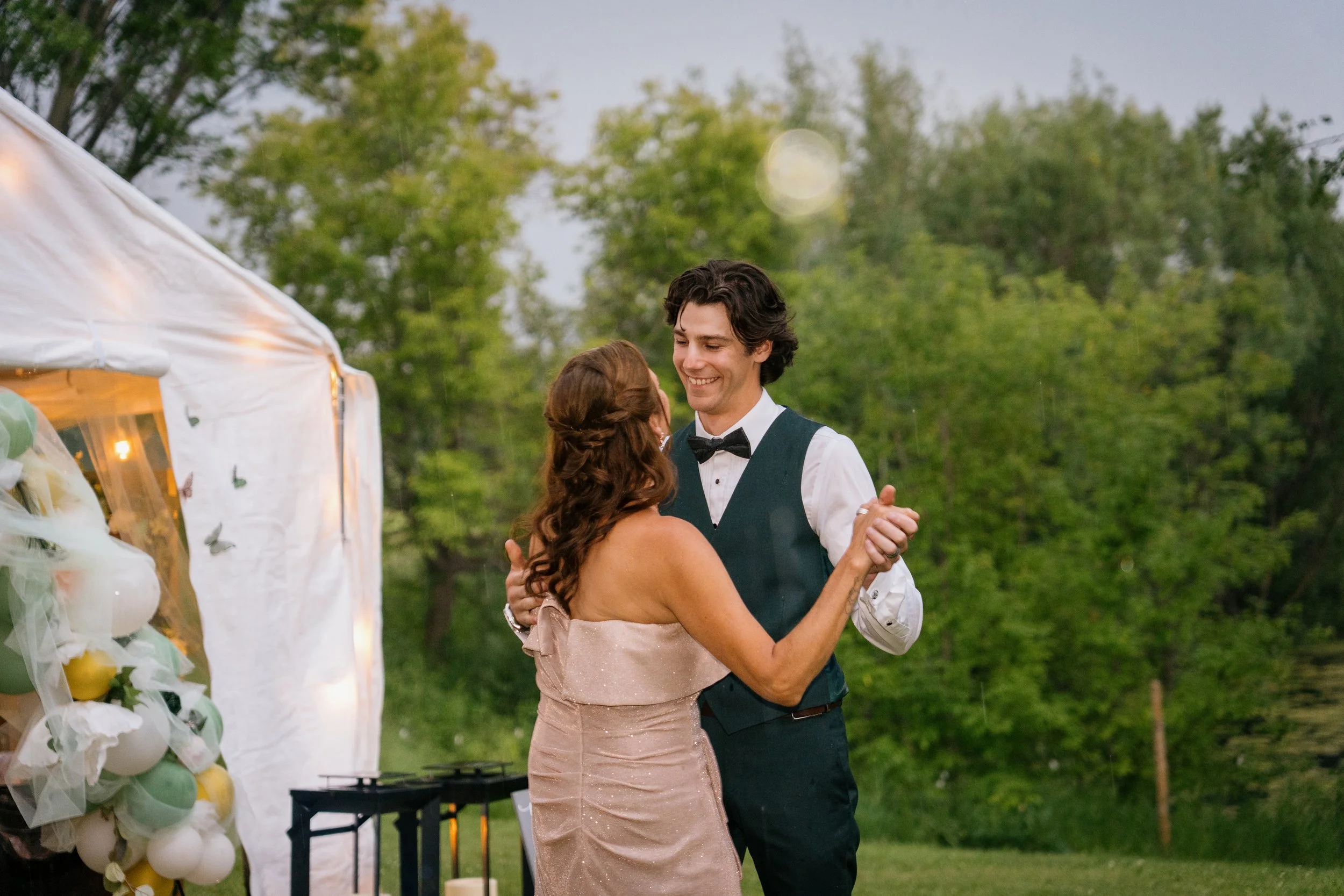 A happy couple dancing outdoors at a wedding reception during the evening, with trees and a decorated balloon arch visible in the background.