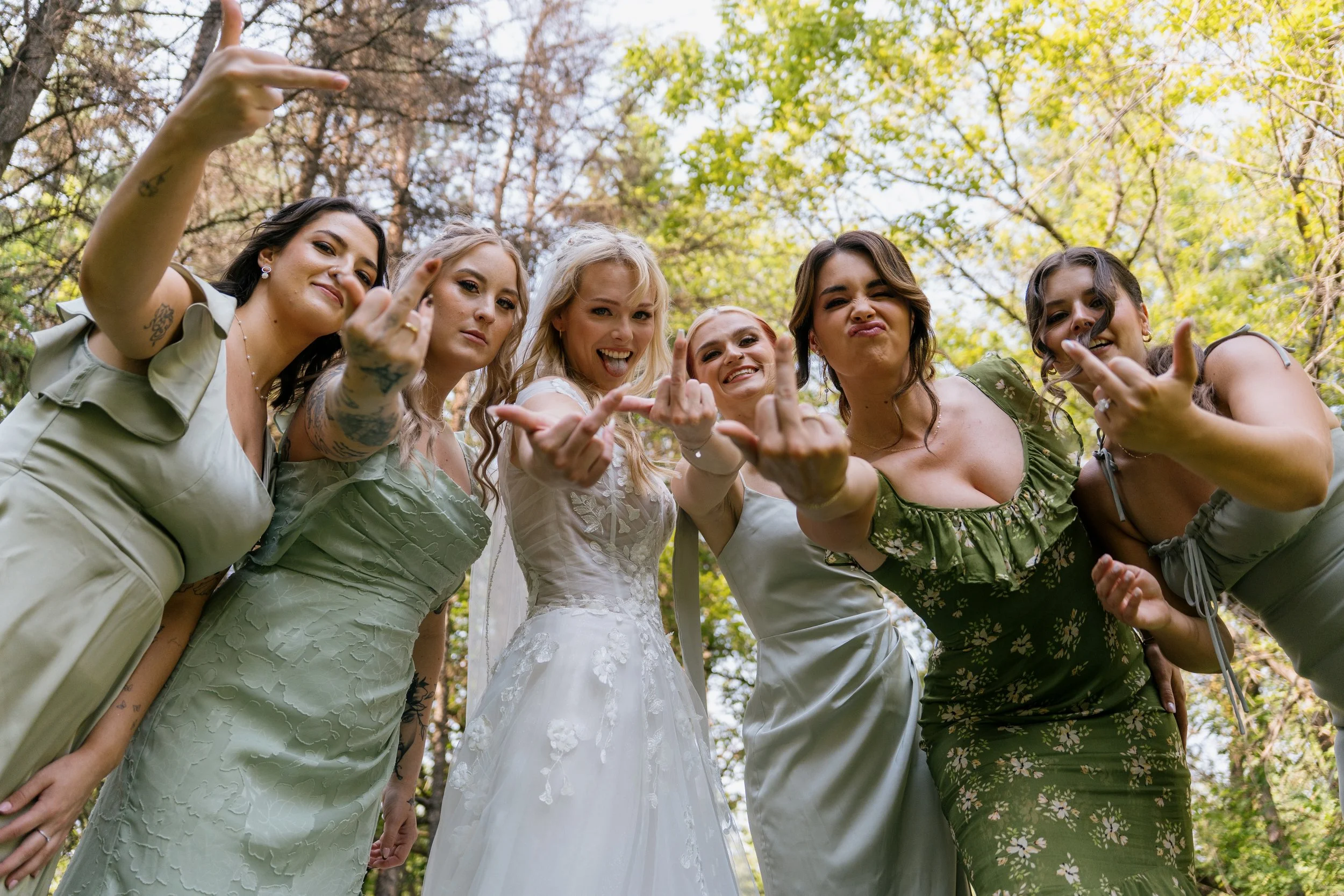 A group of six women standing outdoors in a circle, making hand gestures and smiling, with a background of trees with green and brown leaves.