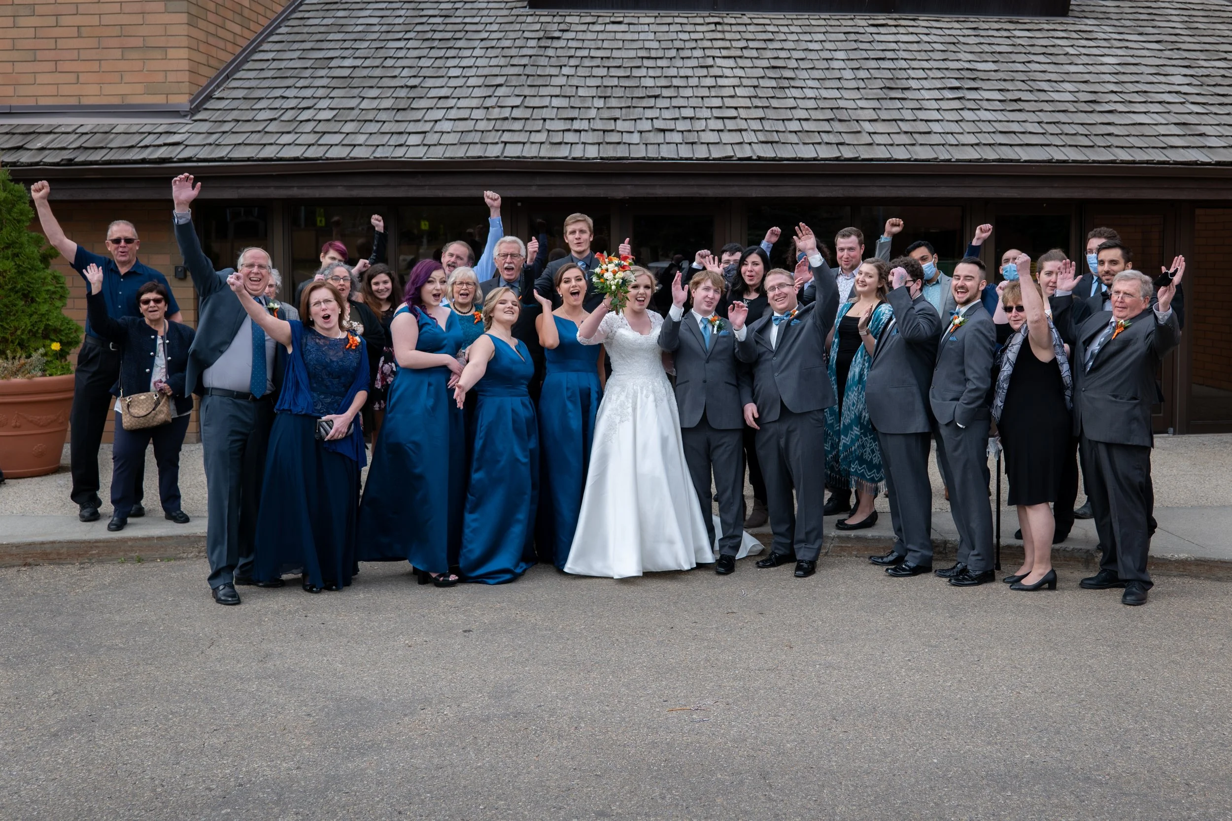 A large group of people celebrating outside a building, with some wearing formal attire, including a bride in a white wedding dress holding a bouquet, and others dressed in suits and dresses, raising their arms in joy.