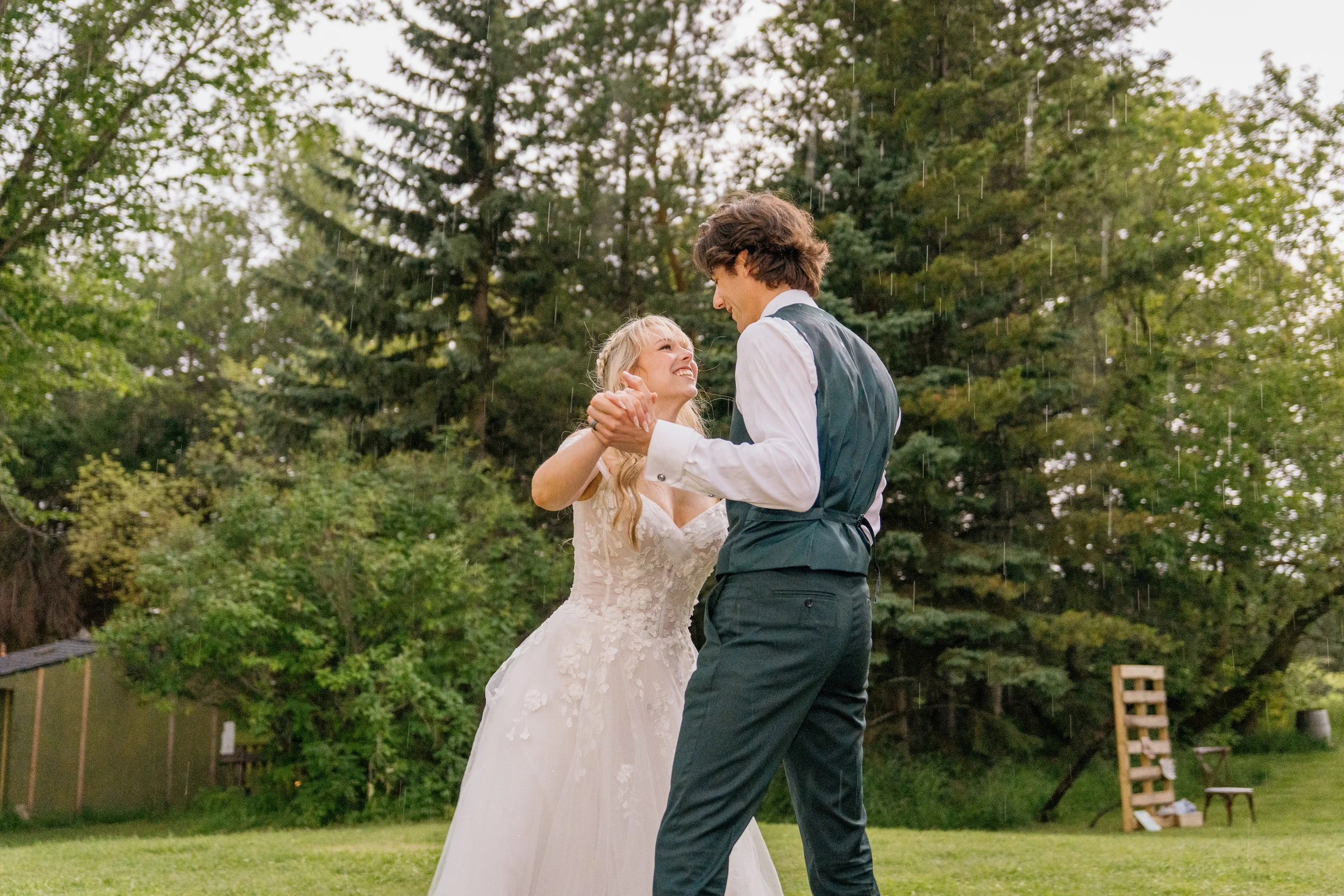A bride and groom dancing outdoors in a garden, surrounded by green trees, on a rainy day.